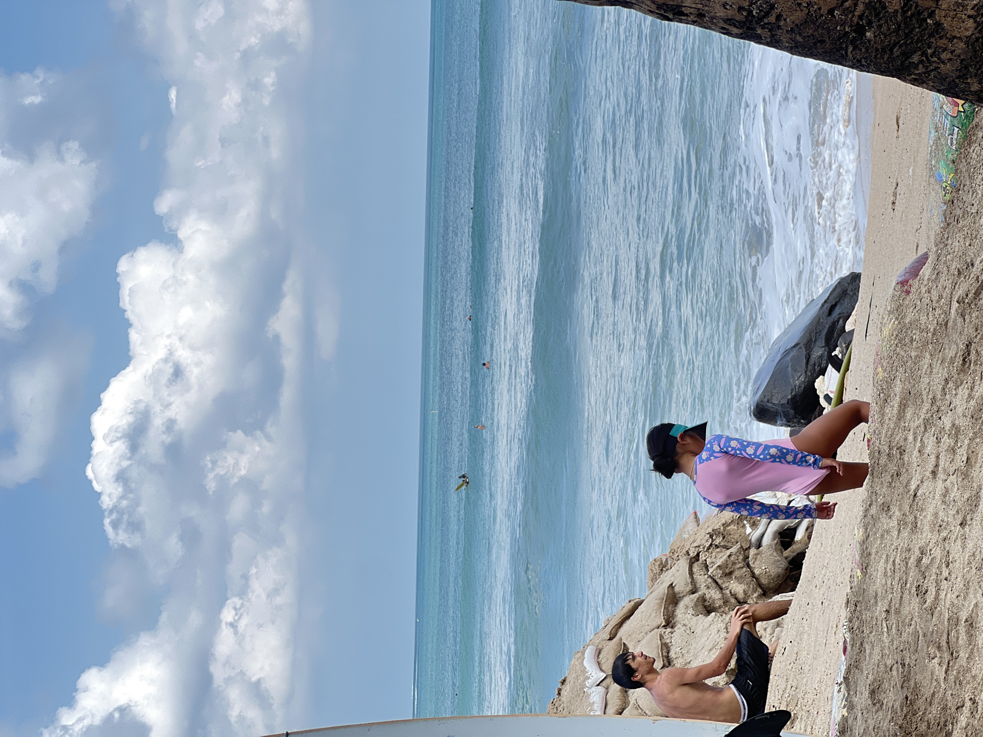 The image depicts a beach scene in Kuta, Indonesia. The sky is a bright blue with fluffy white clouds. The ocean is a vibrant turquoise with gentle waves breaking on the shore.
In the foreground, two people are visible. A young boy is lying on his back on the sand, wearing black shorts and a black cap. His body is partially obscured by a white object, possibly a surfboard. A young girl is sitting on rocks nearby, facing the ocean. She is wearing a pink and blue long-sleeved rash guard and a black visor. She appears to be looking out at the water.
In the ocean, several small figures can be seen, some appearing to be swimming or possibly surfing. The overall atmosphere is one of a relaxed day at the beach. The sunlight suggests it is daytime, likely late morning or early afternoon. The presence of the ocean, sand, and rocks are typical of a beach environment. There is graffiti visible on the rock wall in the far right of the image.