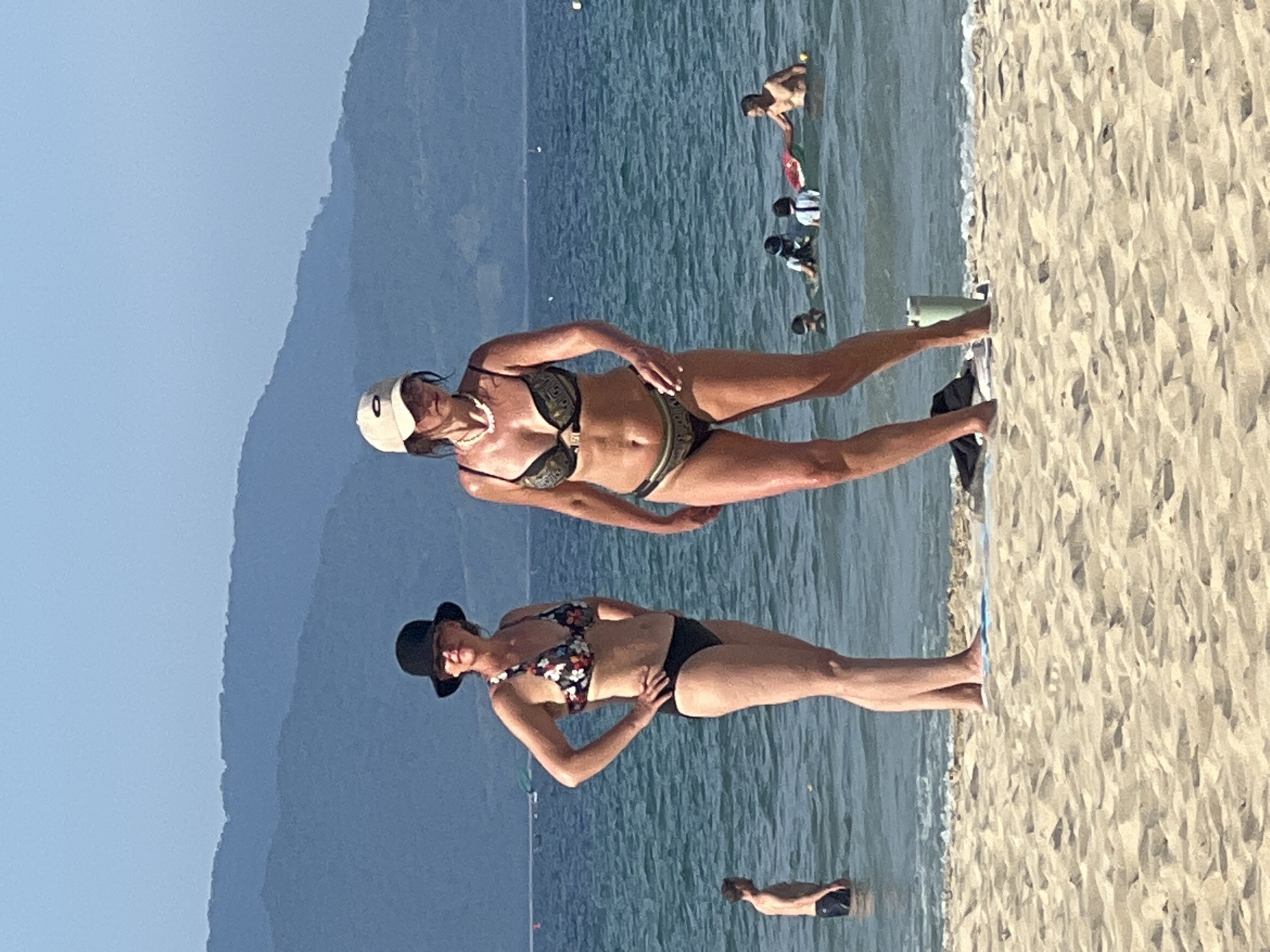 Two women are standing in the shallow water of a beach. The woman on the left is wearing a black hat and a floral bikini. She has her hands on her hips. The woman on the right is wearing a beige baseball cap and a patterned bikini. She is looking down and to her left. In the background, there are mountains and other people in the water. The scene appears to be on a sunny day. The specific location is identified as the city of Đà Nẵng, Vietnam.
