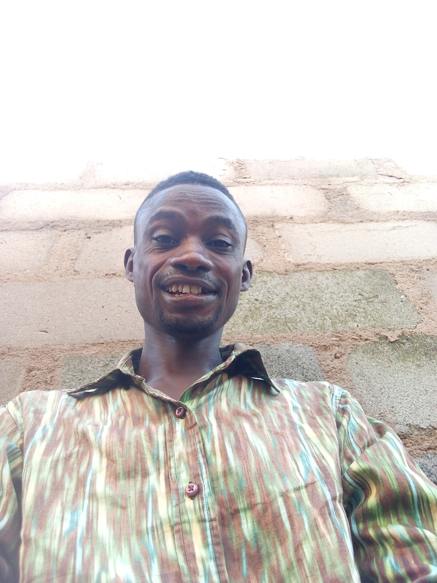 A man with dark skin, short dark hair, and a slight mustache and goatee smiles directly at the camera, showing his upper teeth. He is wearing a patterned button-up shirt with vertical stripes and streaks in shades of green, brown, light blue, and off-white. The shirt has a collar and two visible brown buttons down the front. The image appears to be a selfie taken from a slightly low angle, looking upwards.
The background consists of a rough, light-colored brick or concrete block wall, indicating an outdoor setting. The wall takes up the lower two-thirds of the background, with a brighter, almost white area above, suggesting an overcast sky or bright light source directly overhead. The texture of the wall is prominent, showing individual blocks and mortar lines.
The man's expression is one of warmth and approachability. There is no visible text in the image. The bright lighting suggests it is daytime. Given the location context of Adonkolo, Nigeria, the scene depicts a casual moment in an everyday environment.