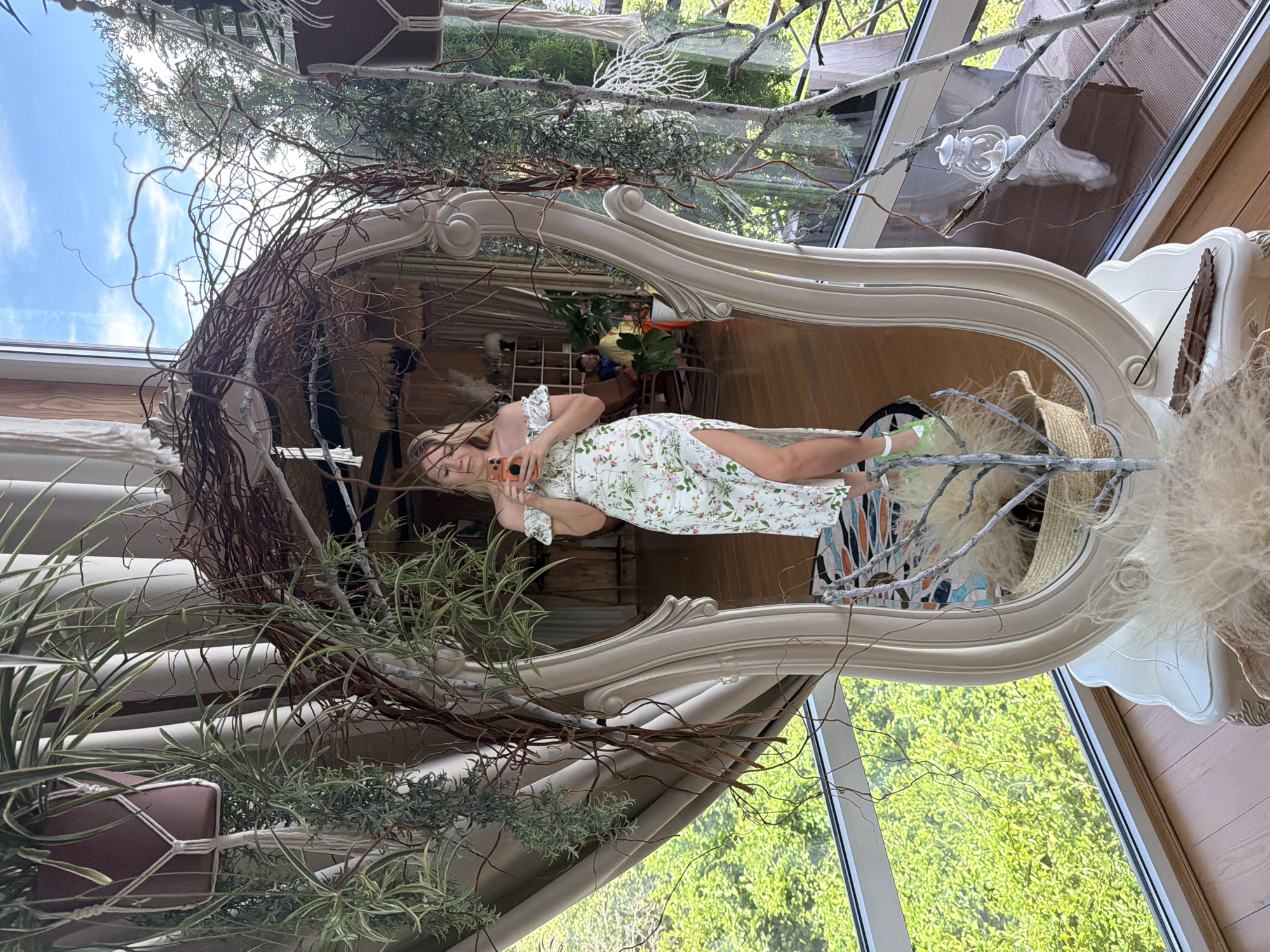 A woman is reflected in a large, ornate mirror, holding a smartphone. She is wearing a white floral off-the-shoulder dress with a high slit and green sneakers. The mirror is framed by hanging plants and dried branches, creating a bohemian atmosphere. Behind her, through large windows, lush green foliage is visible, suggesting an outdoor setting. The interior decor appears to be eclectic, with macrame plant hangers and a wicker basket. The scene is captured indoors, likely in a home or a studio, with the reflection in the mirror showing more of the room's interior and a hint of an outdoor view. The location is specified as Nizhnyaya Shilovka, Russia.