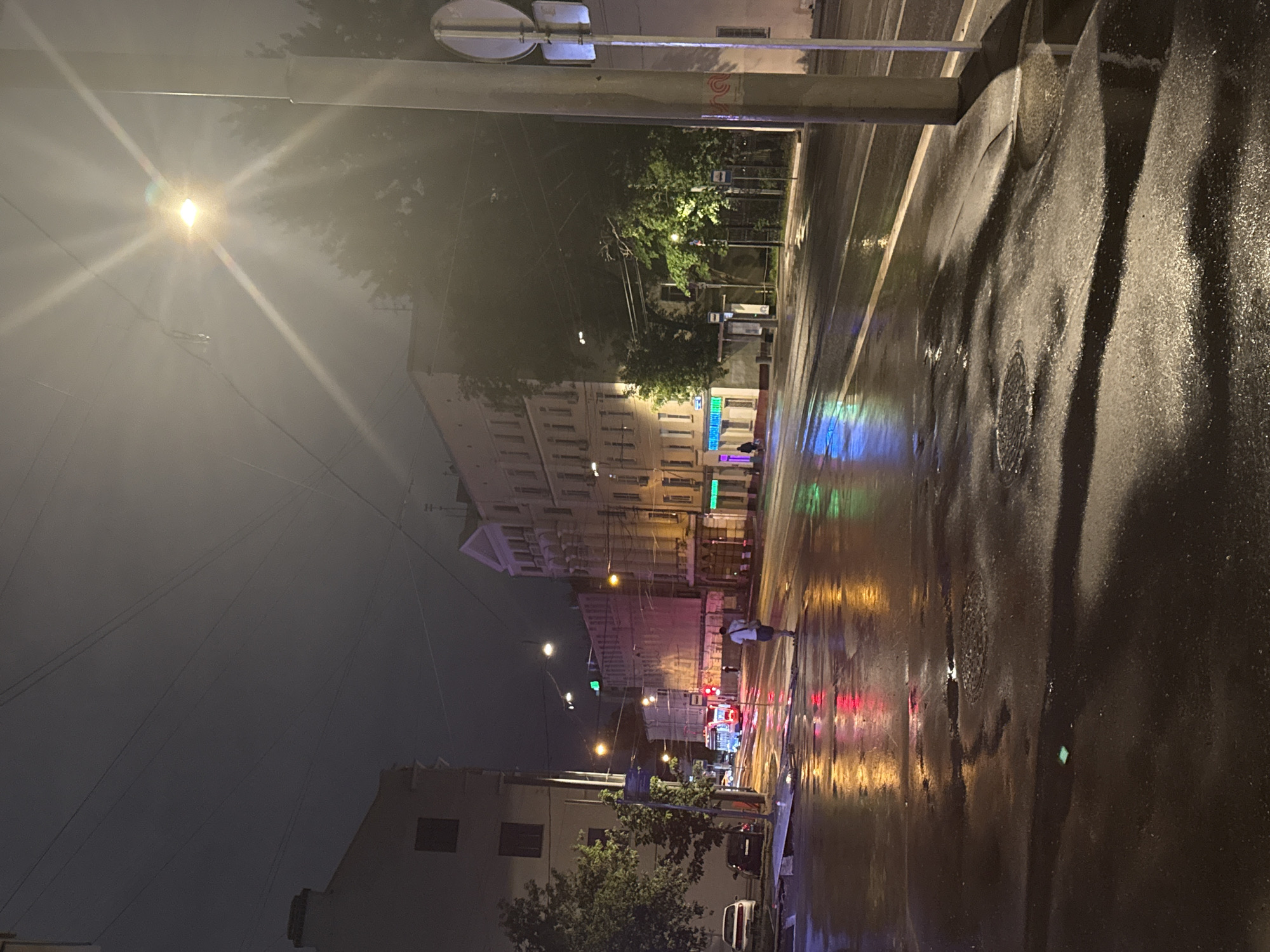 The image depicts a street scene in Moscow, Russia, at night, following rainfall. The foreground shows a wet asphalt surface reflecting the surrounding lights. A street lamp with a noticeable lens flare is visible on the left. Buildings line the street, with their facades illuminated by streetlights and neon signs. In the distance, car headlights and taillights create streaks of red and white. A person is walking along the sidewalk on the right side of the road. Trees are interspersed along the street, adding to the urban landscape. A street sign is visible in the top left corner.