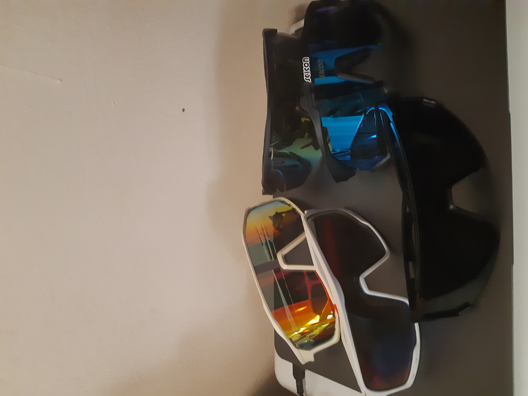 The image shows a pair of ski goggles and two interchangeable lenses mounted on a dark grey wall panel with the brand name "scicon" visible. The lenses are reflective and show a blue and orange hue. The wall appears to be off-white. The scene is likely indoors. The location is not evident in the image and cannot be determined.