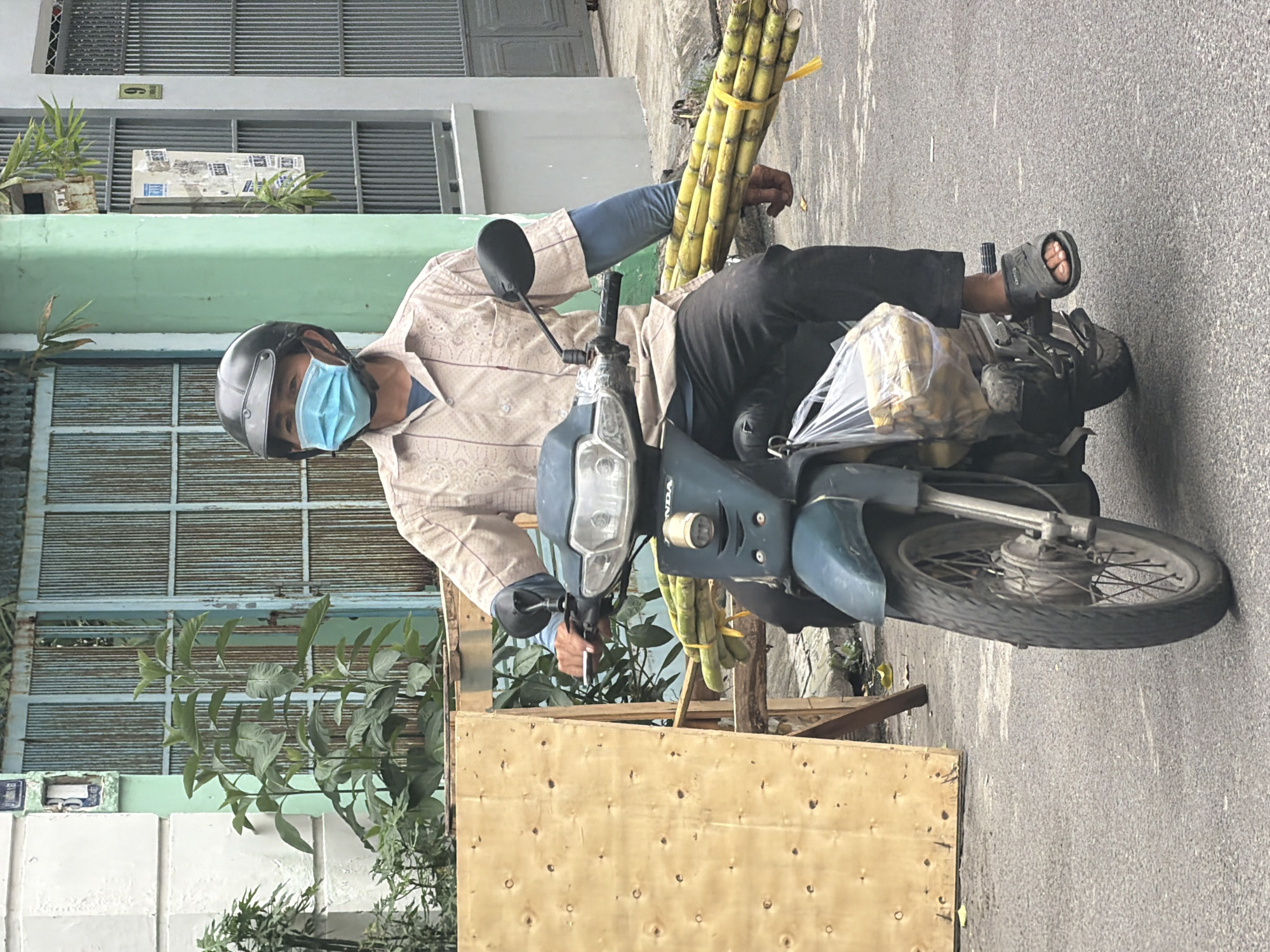 In Nha Trang, Vietnam, a person wearing a light-colored patterned shirt, black pants, a black helmet, and a light blue face mask rides a dark green scooter. The person is carrying a large bundle of sugarcane stalks, tied with yellow twine, on the right side of the scooter. A clear plastic bag filled with what appears to be yellow food items is hanging from the left side of the scooter. The scooter is on an asphalt road. In the background, there is a light green building, a large wooden panel, and a metal gate with vertical bars.