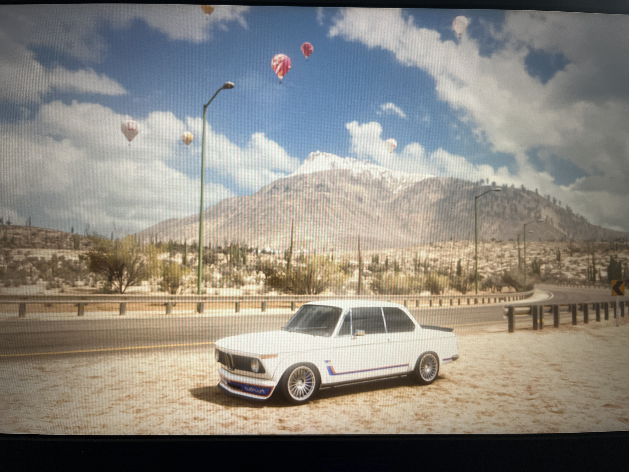 The image depicts a desert landscape with a white vintage BMW 2002 car parked on a dirt road. The car has blue and red racing stripes along its side and a spoiler with blue and red accents. In the background, a mountain range with snow-capped peaks rises against a bright blue sky dotted with fluffy white clouds. Several hot air balloons of various colors, including red, white, and yellow, are visible in the sky. The terrain is arid, with sparse desert vegetation like cacti and shrubs. A road with a guardrail runs alongside the car. Streetlights are placed along the road. The scene appears to be set during the daytime, with clear skies and bright sunlight, suggesting fair weather. There is no visible text in the image, except for the word "SOUL" printed on the front bumper of the car. The overall impression is a scenic and somewhat surreal setting, possibly from a video game. The prompt specifies Varna, Bulgaria as a location context, but the visual elements strongly suggest a desert environment, not typical of Varna.
