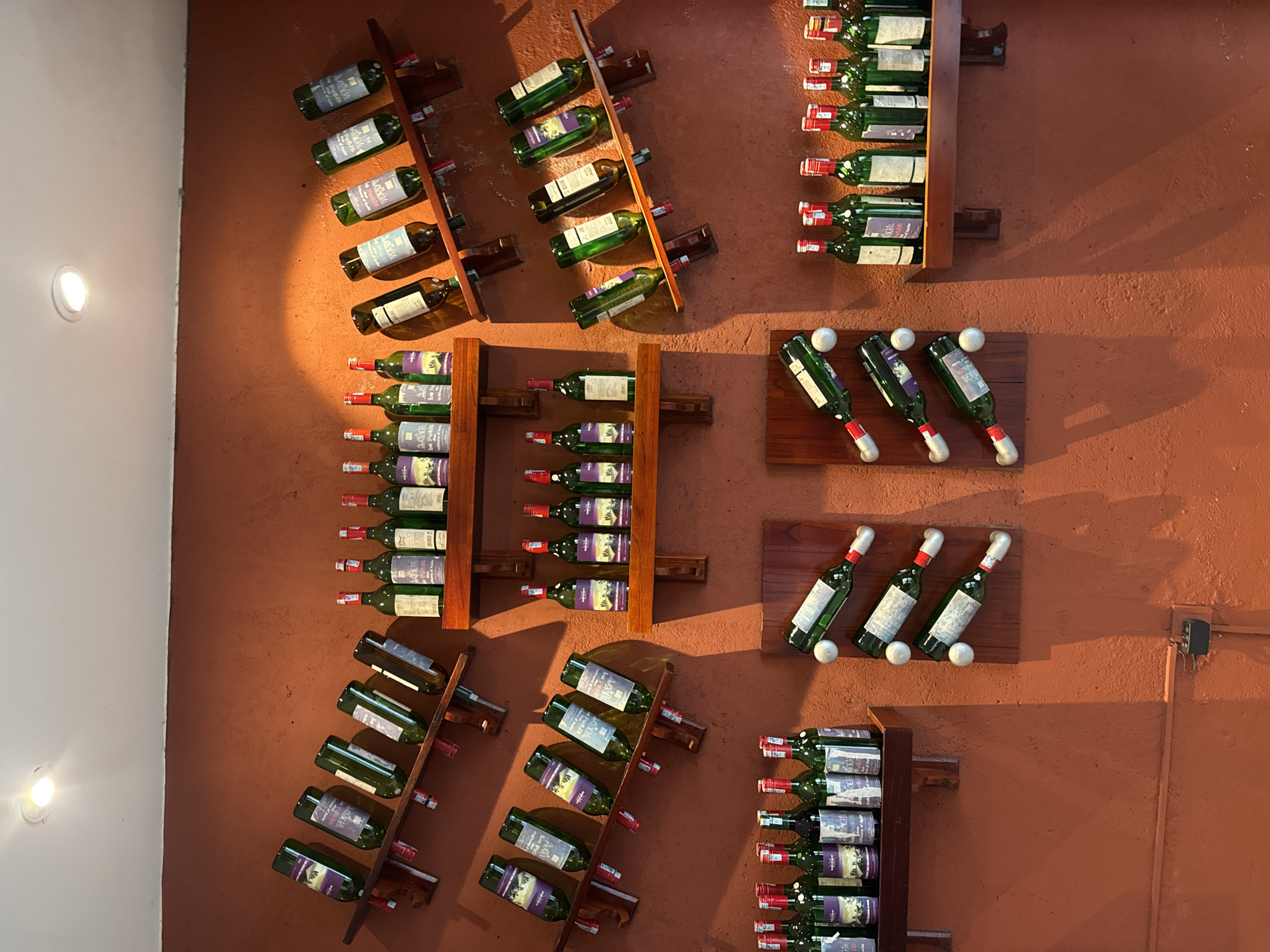 The image displays an indoor wall decorated with a large collection of wine bottles, predominantly in green glass, showcased on various wooden racks. The wall itself is a reddish-brown with a textured surface. On the far left, a smooth white wall section features two circular, flush-mounted white lights.
The wine bottles are arranged in several distinct patterns:
- In the upper left and middle right sections, bottles are displayed on angled wooden racks, often tilted downwards.
- In the upper right section, bottles are arranged horizontally on shorter wooden shelves.
- In the middle and lower left, and lower right sections, tall, vertical wooden racks hold multiple bottles in stacked or side-by-side configurations.
- In the middle right, several bottles are uniquely presented horizontally on wooden plaques, held in place by spherical white objects resembling golf balls that secure the bottle necks and bases.
Many of the wine labels are visible, and common text includes "DALAT" and "Vang Đàlạt," indicating Vietnamese Dalat Wine. Other legible words on labels include "Classic" and "Export."
Artificial lighting, likely emanating from the lights on the white wall and possibly other hidden sources, casts distinct shadows from the bottles and racks onto the textured wall, suggesting an indoor setting with directed illumination. A small, black electrical box is mounted on the wall in the lower right. The overall scene creates a rustic yet organized wine display, located in Đà Nẵng, Vietnam.