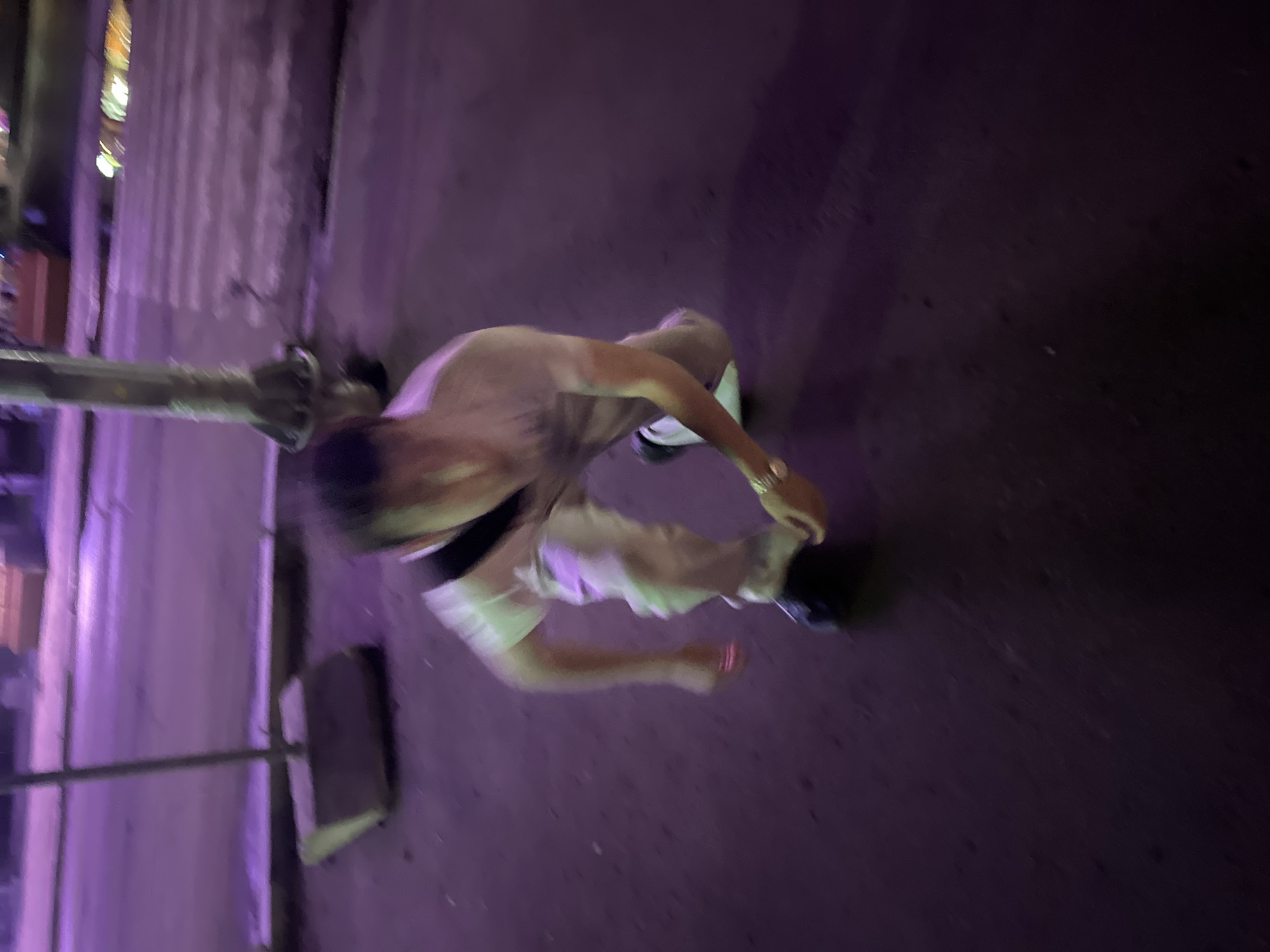 The image shows a person with short, dark hair and fair skin, wearing a light-colored sleeveless shirt and light-colored shorts with a dark waistband. They appear to be standing on a street at night, with the ground illuminated by purple lights. The person is leaning forward, with their left arm extended and their right hand near their waist. A metal pole is visible in the foreground, extending from the left side of the image. The background is blurred and indistinct, but appears to be a street with some building facades and lights. The overall impression is that of a candid street photography shot taken at night in an urban environment. The lighting suggests it is nighttime. There is no visible text in the image.
