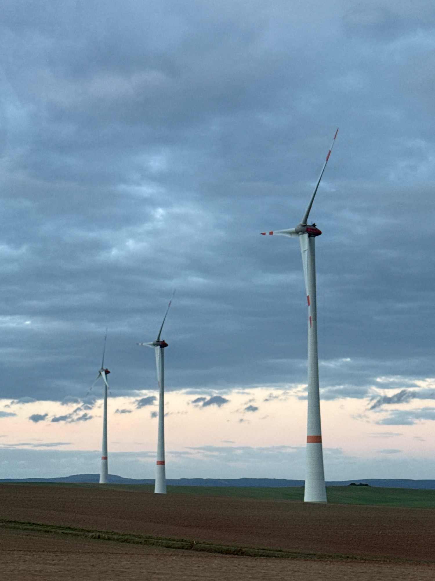 Three wind turbines stand in a field under a cloudy sky. The turbines are white with orange bands and red tips on their blades. The closest turbine is on the right and is fully visible. The other two turbines are to the left and are partially obscured by the closest one and by distance. The ground in the foreground is brown and appears to be cultivated. In the background, there are rolling green hills. The sky is filled with dark, heavy clouds, with a sliver of lighter sky visible near the horizon. The image appears to have been taken at dusk or dawn due to the lighting and sky conditions. This scene is typical of the countryside surrounding Sondershausen, Germany, a region known for its wind farms. There are no people or animals visible in the image.