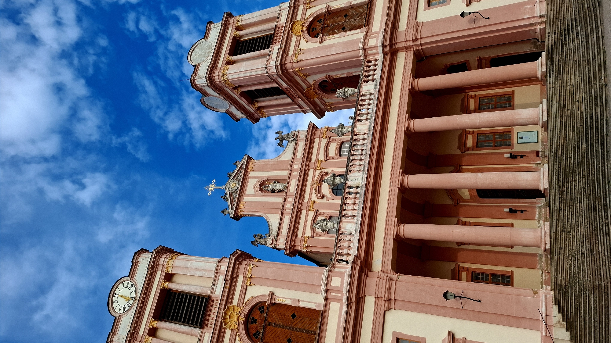 The image shows the exterior of the Göttweig Abbey, a Benedictine monastery located near Furth bei Göttweig, Austria. The photograph is taken from a low angle, looking up at the facade of the abbey church, which is characterized by its pink and cream-colored Baroque architecture.
The facade features two prominent bell towers flanking a central section topped with a cross. Both towers have clocks with Roman numerals and arched windows. The central part of the church has intricate sculptural details and a large balcony supported by columns. Below the balcony, a series of large, pink columns support the upper structure of the building. A wide, stone staircase leads up to the entrance.
The sky is a vibrant blue with scattered white clouds, suggesting it is daytime and the weather is fair. There are no people visible in the frame. The overall impression is one of grandeur and historical significance.