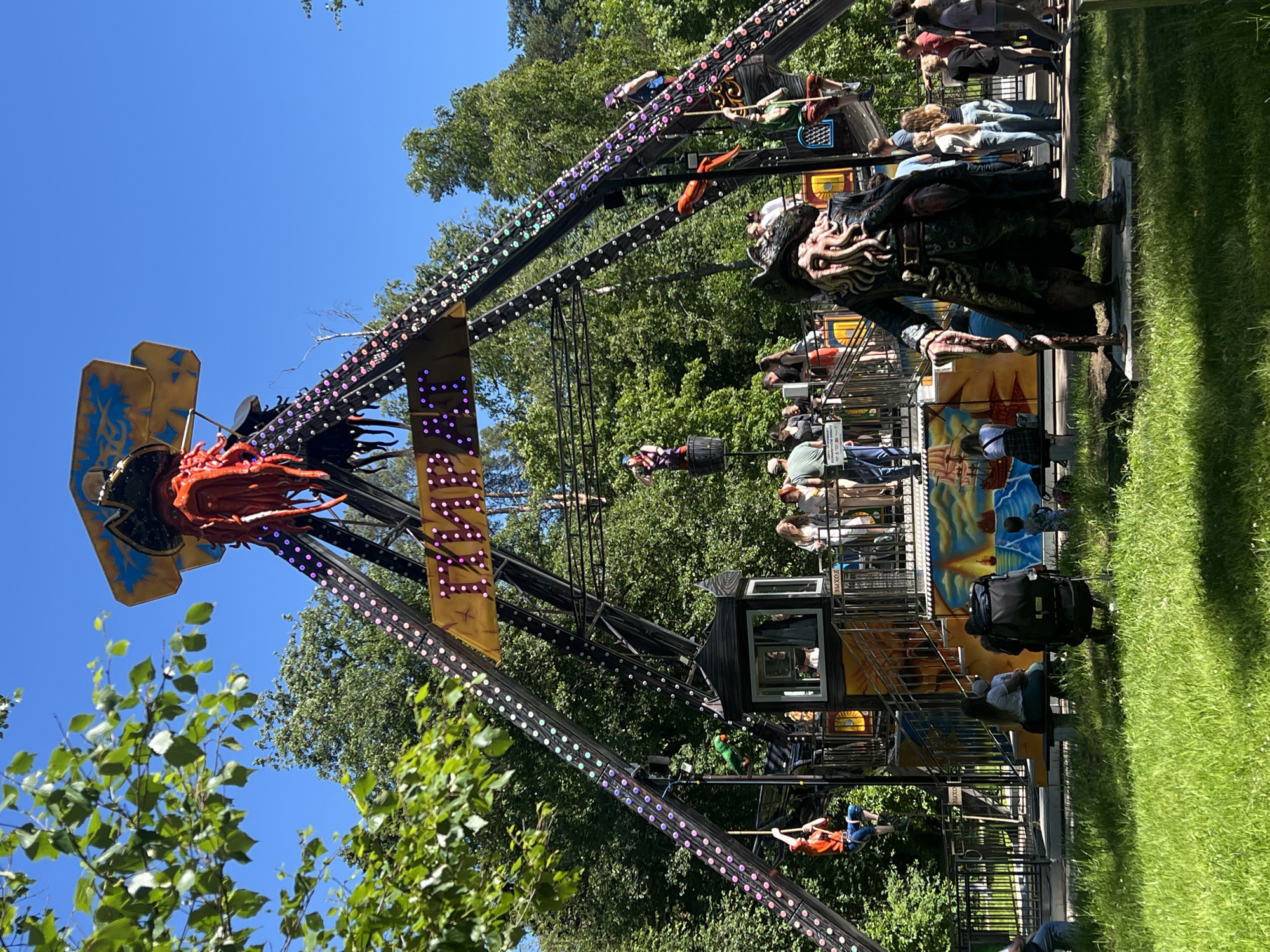 This is a medium shot, outdoor photograph taken on a sunny day at an amusement park in Zelenogorsk, Russia. The focal point of the image is a large, pirate-themed amusement ride with a pendulum-like structure. The ride is adorned with numerous colorful lights and features a prominent pirate skull and tentacles at the top. A banner with Cyrillic text and lights hangs across the structure.
In the foreground, a statue of a pirate with a long beard and tentacles for a face stands on a concrete base, facing the ride. To the left and right of the ride, various people are visible. Some are waiting in line, others are riding the attraction, and some are standing and observing. Children are also present, with one child sitting on a bench with an adult and another child walking near a stroller. The environment is lush and green, with grass in the foreground and mature trees in the background, indicating a park or outdoor recreational area. The sky is clear and blue.