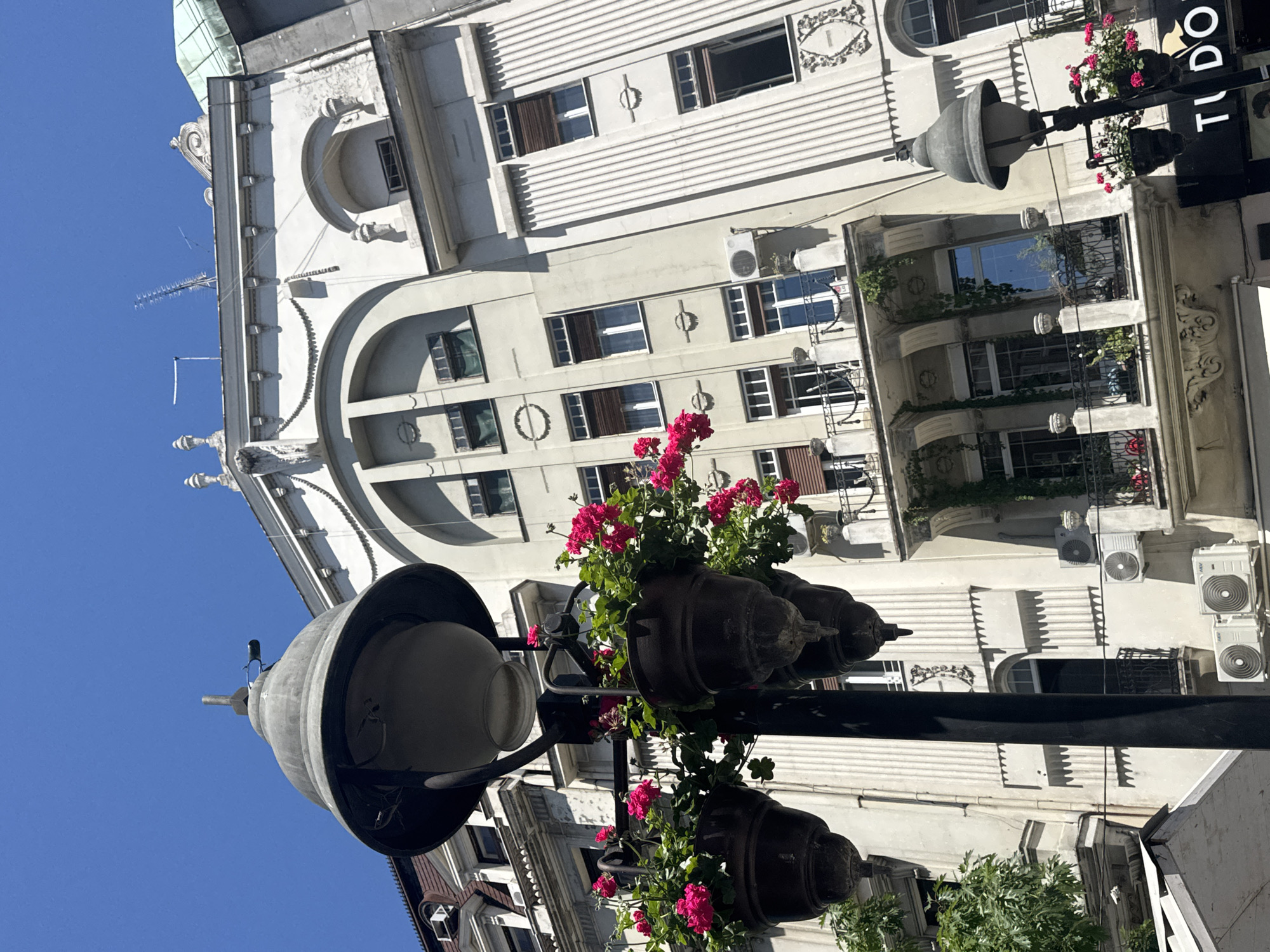 The image depicts a street scene in Belgrade, Serbia, dominated by a vintage lamppost adorned with hanging flower baskets filled with bright pink flowers. Behind the lamppost, an ornate, light-colored building with a curved facade and arched windows stands tall. Architectural details such as balconies with wrought-iron railings, decorative moldings, and classical ornamentation are visible on the building's exterior. The sky above is a clear, bright blue. Several air conditioning units are mounted on the building's facade. A black and white sign with the text "TUDOR" is partially visible at the bottom right of the image. The overall impression is one of a charming, sunlit urban environment.