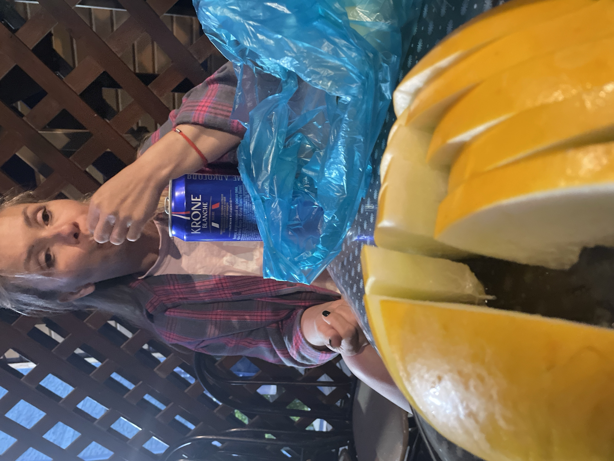 A medium shot shows a woman eating with her right hand in the foreground, to the left of a blue plastic bag. The woman has grey hair and wears a pink and grey plaid shirt over a light pink t-shirt. She has a red string bracelet on her left wrist. Next to her is a blue can of Krone Blanche beer. In the foreground of the image are slices of yellow melon arranged on a dark surface. The background shows a wooden lattice fence. The image is an outdoor scene, possibly on a patio, with a glimpse of blue water visible through the fence.