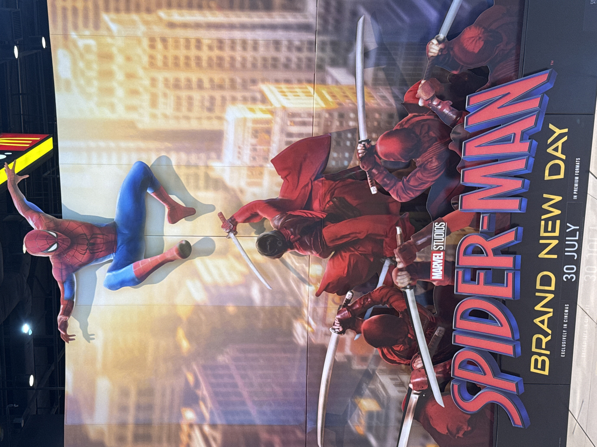 This is a vertical, eye-level, medium shot of a promotional display for "Spider-Man: Brand New Day." The display features a life-size cutout of Spider-Man in his iconic red and blue suit, posed mid-leap on the left side. To the right, several figures dressed in red, resembling Daredevil, are depicted in action poses, wielding swords. The background is a blurred cityscape with a warm, golden hue, suggesting a sunset or sunrise.
The dominant text in the image reads "SPIDER-MAN BRAND NEW DAY" in large, embossed letters. Below this, "MARVEL STUDIOS" is visible, along with "30 JULY" and "EXCLUSIVELY IN CINEMAS," indicating the film's release date and where it can be seen. The phrase "IN PREMIUM FORMATS" is also present.
The image appears to have been taken indoors, possibly in a cinema lobby or a retail space, given the lighting and the backdrop. The overall mood is action-oriented and exciting, reflecting the superhero theme. The "Bangkok, Thailand" context suggests this promotional material was displayed in that city.