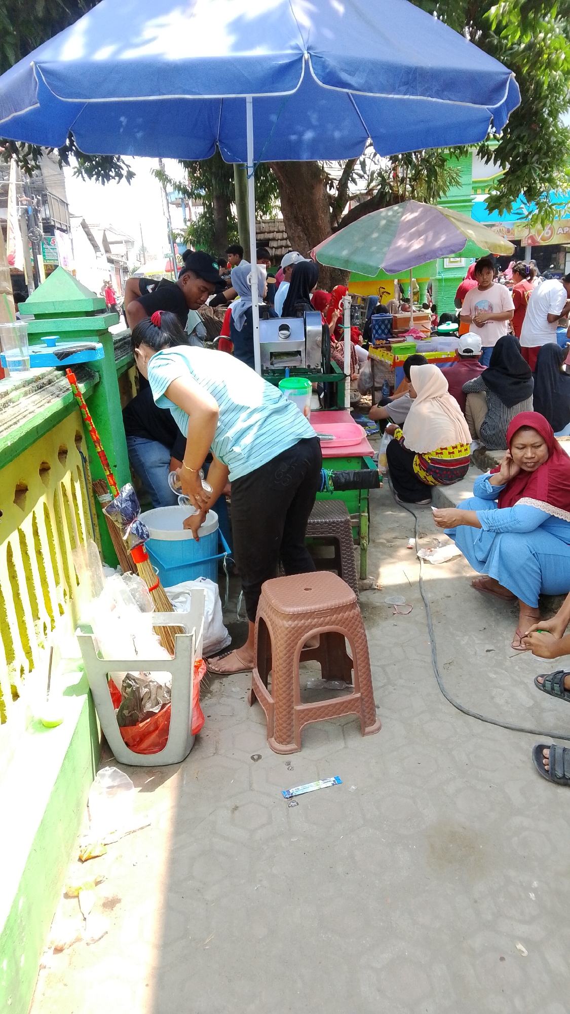 The image shows an outdoor market scene in Mancar, Indonesia, with several people gathered under a large blue umbrella. In the foreground, a woman wearing a blue and white striped shirt and black pants is leaning over a blue bucket, appearing to be washing her hands or an object. To her left, a broom is leaning against a green and yellow wall. Several plastic bags and a white plastic container are also visible near the wall.

In the middle ground, a sugar cane juicing machine is set up, with several individuals around it. Some people are seated on the ground or on small stools. A woman in a red hijab and blue outfit is sitting and talking on a cell phone. Another woman is wearing a light pink hijab.

The background shows more market stalls and trees, with a green building visible in the distance. The ground is paved with concrete. The overall atmosphere appears to be busy and informal.