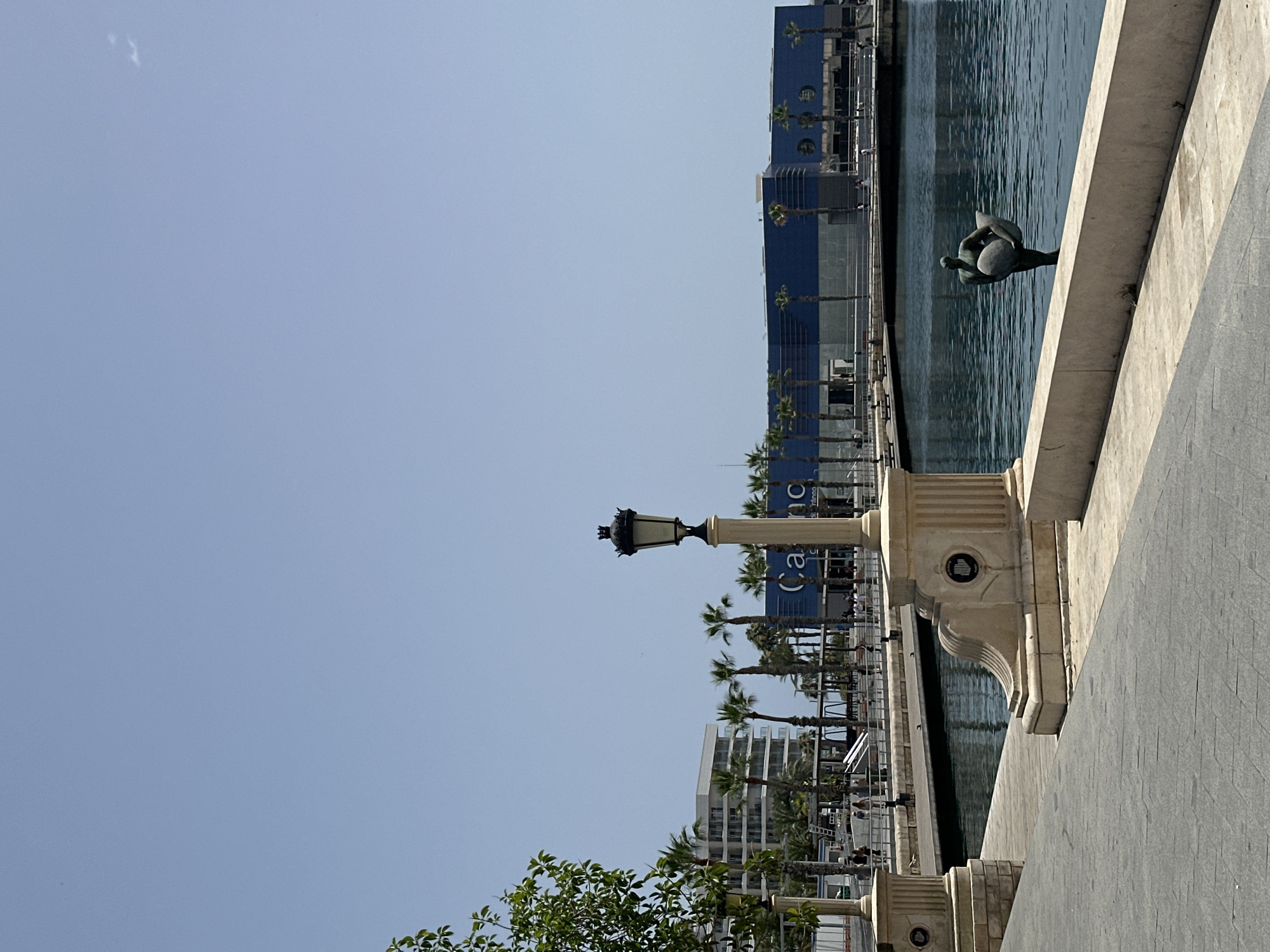 A bright, sunny day is captured in this scene along a waterfront promenade. The foreground is dominated by a paved walkway with a textured gray surface, leading the eye towards the body of water. To the left, a light-colored stone pillar with decorative carvings stands tall, topped with an ornate black lamppost. A similar pillar is visible further down the walkway.
The calm, blue water of the harbor is on the right, reflecting the clear sky and the surrounding structures. In the water, a bronze statue depicts a human figure holding a large, oval-shaped object. A low, cream-colored wall separates the walkway from the water.
In the mid-ground, a wooden boardwalk runs parallel to the water, with people strolling along it. Beyond the boardwalk, a modern, white building with multiple balconies rises, surrounded by palm trees. Further in the background, a large, blue building with a modern facade is visible, featuring large circular windows and the word "Casino" prominently displayed on its exterior. More palm trees line the waterfront in front of this building. The sky above is a clear, pale blue with a few wispy clouds.