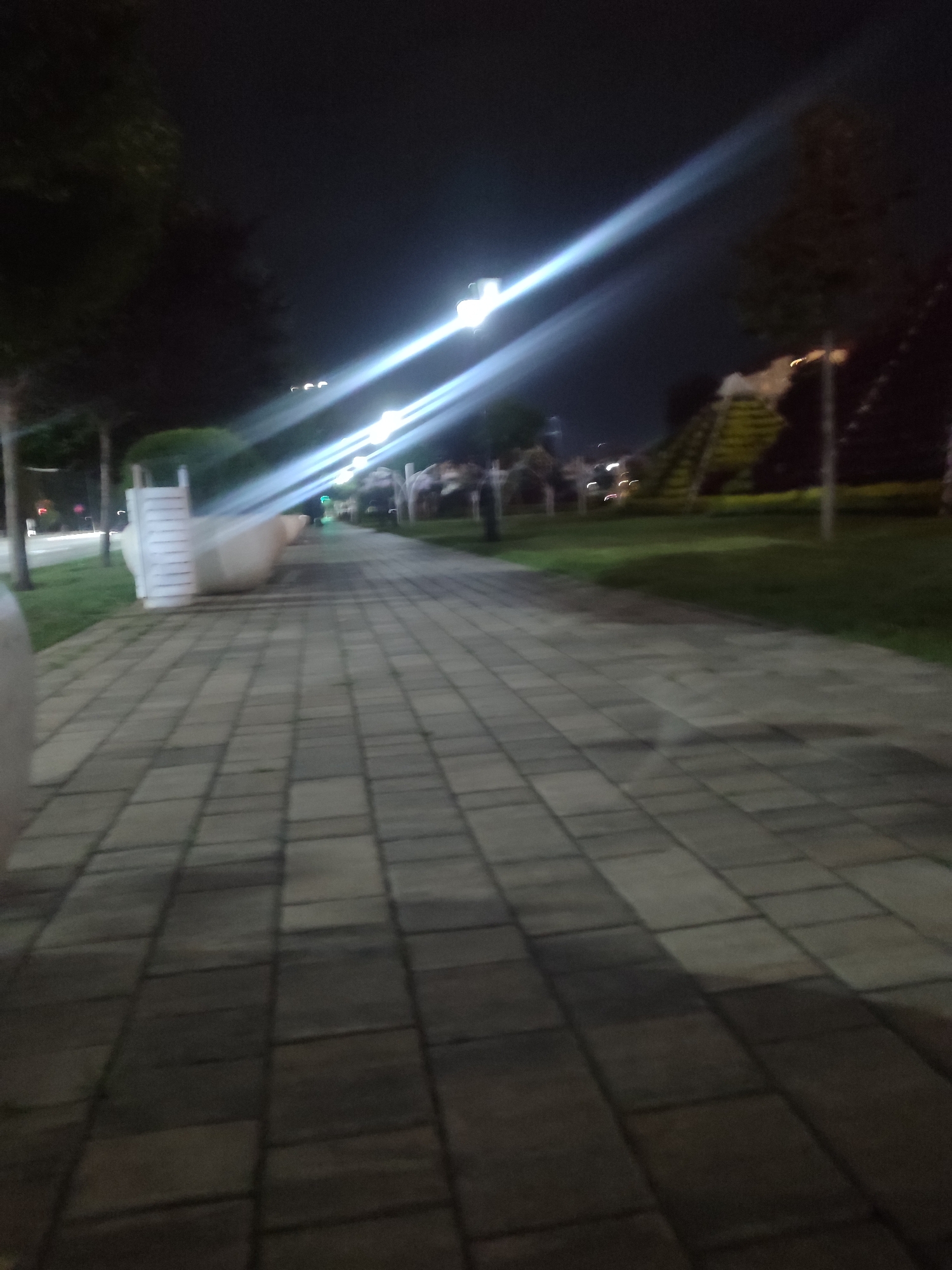 The image depicts a nighttime scene of a paved pedestrian walkway in a park-like setting. The walkway is constructed of rectangular stone pavers laid in a grid pattern. To the left of the walkway, there is a patch of grass and trees. Further down the walkway, on the right side, there is more grass and what appears to be a tiered, landscaped structure, possibly a small garden or decorative element. In the background, several streetlights illuminate the scene, casting long streaks of light across the image due to the long exposure of the photograph. The overall impression is of a well-maintained public space at night. The image does not contain any identifiable people or specific landmarks that would allow for precise location identification within Grozny, Russia.