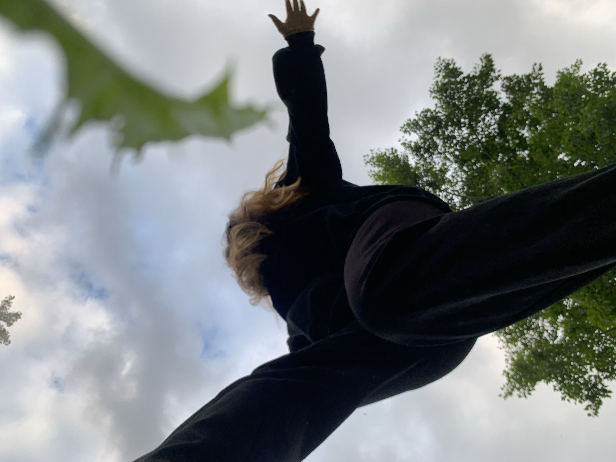 The low-angle, upward-facing perspective shot captures a person in black clothing, seemingly standing or leaping outdoors against a backdrop of cloudy sky and lush green trees. The person's body is mostly silhouetted, with their hair visible as blonde, and one arm extended outwards. A blurred green leaf is also in the foreground, partially obscuring the sky. The image appears to have been taken in the city of Chaikovsky, Russia, as indicated by the context provided.