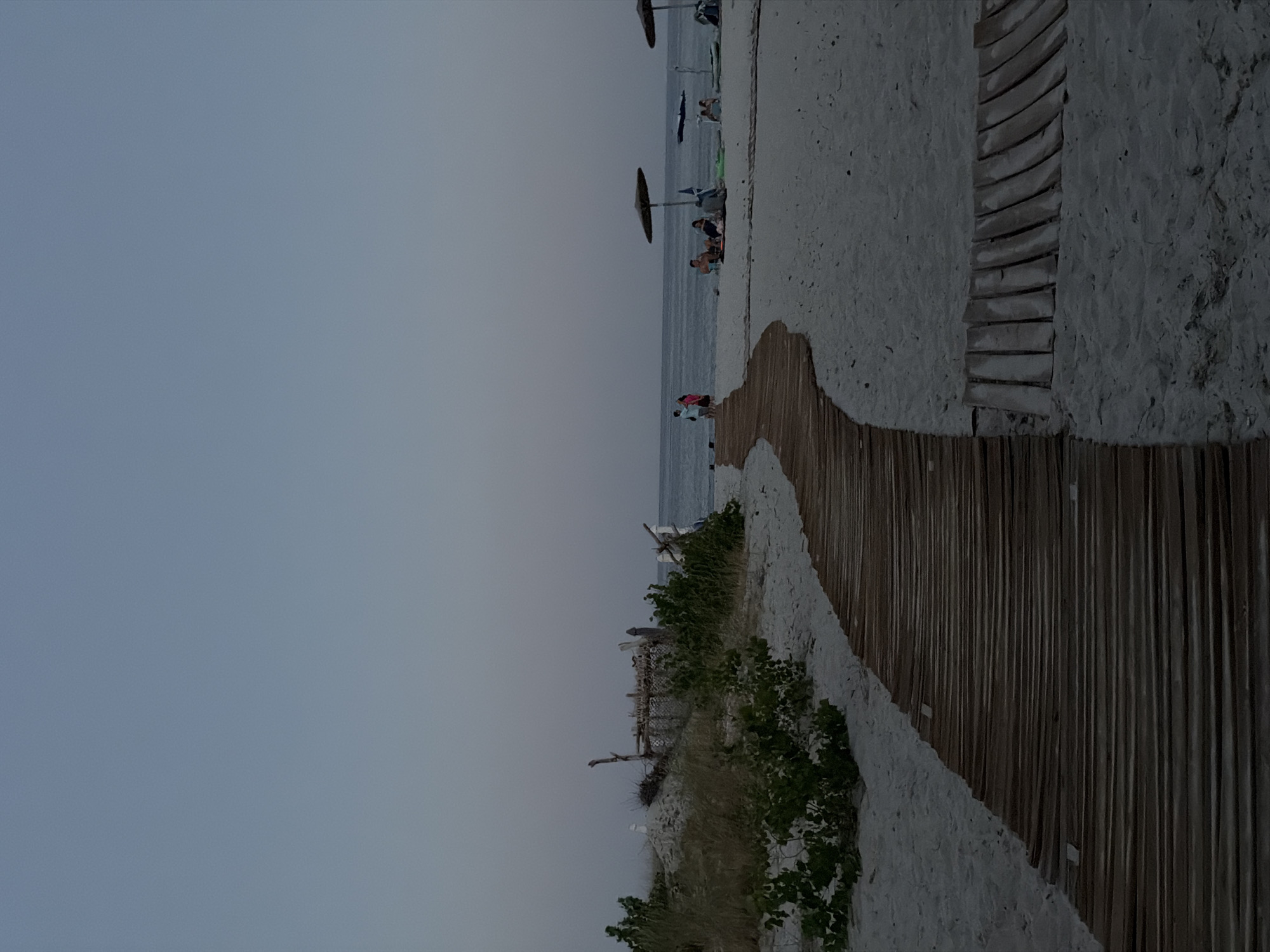 This image depicts a tranquil scene at Golden Beach, Greece, during the late evening. A weathered wooden boardwalk curves across the sandy beach, leading towards the gentle waves of the Aegean Sea. To the left, sparse vegetation and remnants of a makeshift structure suggest a natural, untouched area of the beach. Further down the coastline, several groups of people are scattered, some seated under umbrellas, enjoying the calm atmosphere. The sky is a muted gradient of blue and grey, indicating the twilight hour. The overall impression is one of peaceful relaxation and the serene beauty of a coastal evening.