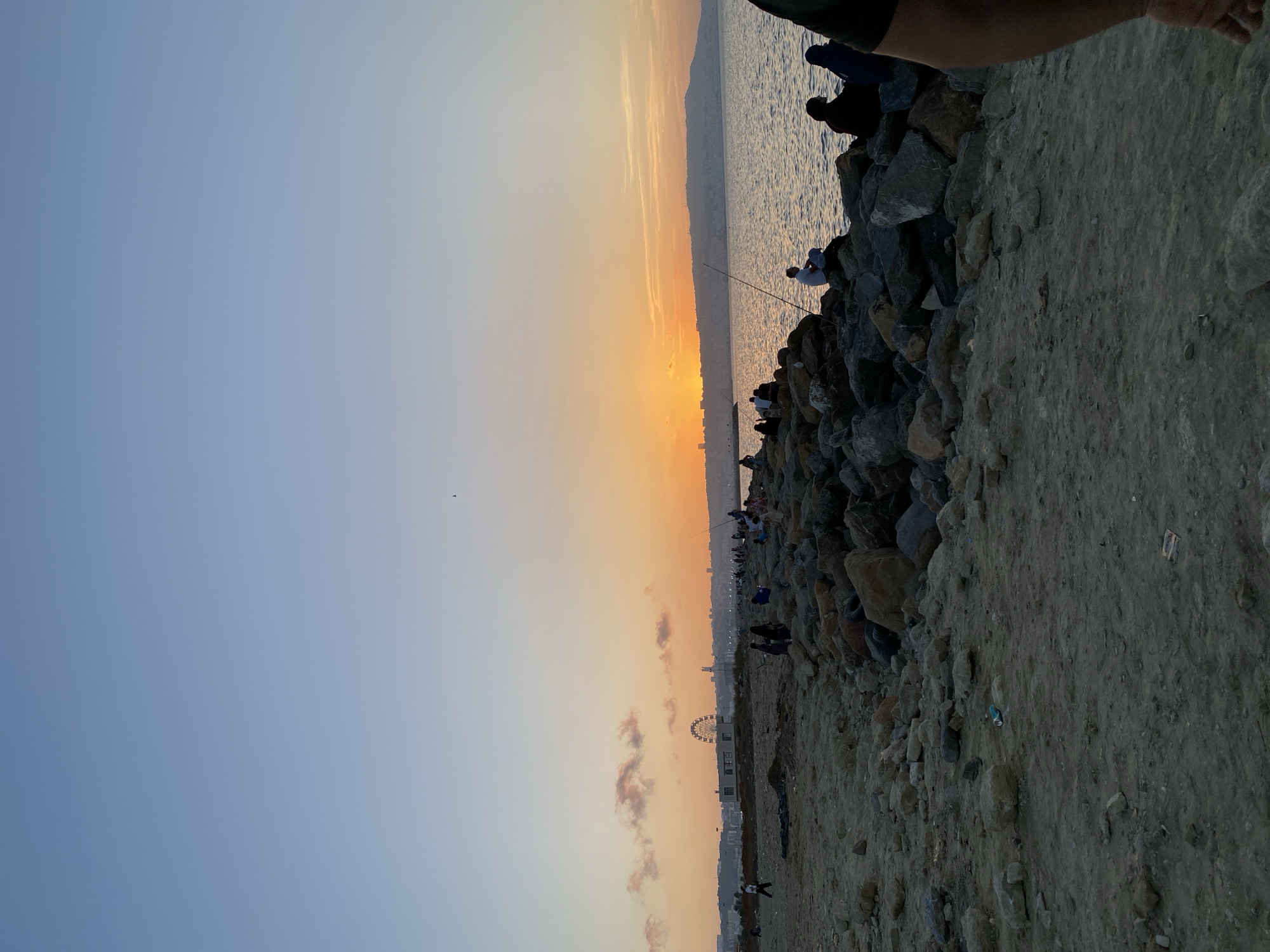 A wide-angle, eye-level, outdoor photograph captures a sunset over the sea from a rocky, sandy shoreline in Hussein Dey, Algeria. The sky transitions from a clear, pale blue at the top to a gradient of orange and yellow near the horizon, where the sun is setting. A thin line of clouds is visible near the sunset. The sea is calm, with gentle ripples reflecting the sunset colors.
On the left side of the frame, a sandy and rocky area extends towards the water, with some scattered debris visible. Further inland, a Ferris wheel and some buildings form part of the cityscape. The right side of the frame is dominated by a rocky breakwater where several people are gathered. Some are seated on the rocks, while others appear to be fishing. One individual in the foreground on the far right has their arm visible, cropped from the frame.
The overall scene depicts a popular recreational area, likely a promenade or fishing spot, where people are enjoying the evening by the sea during a sunset. The location is identifiable as Hussein Dey, Algeria, based on the distinctive Ferris wheel and coastal landscape.