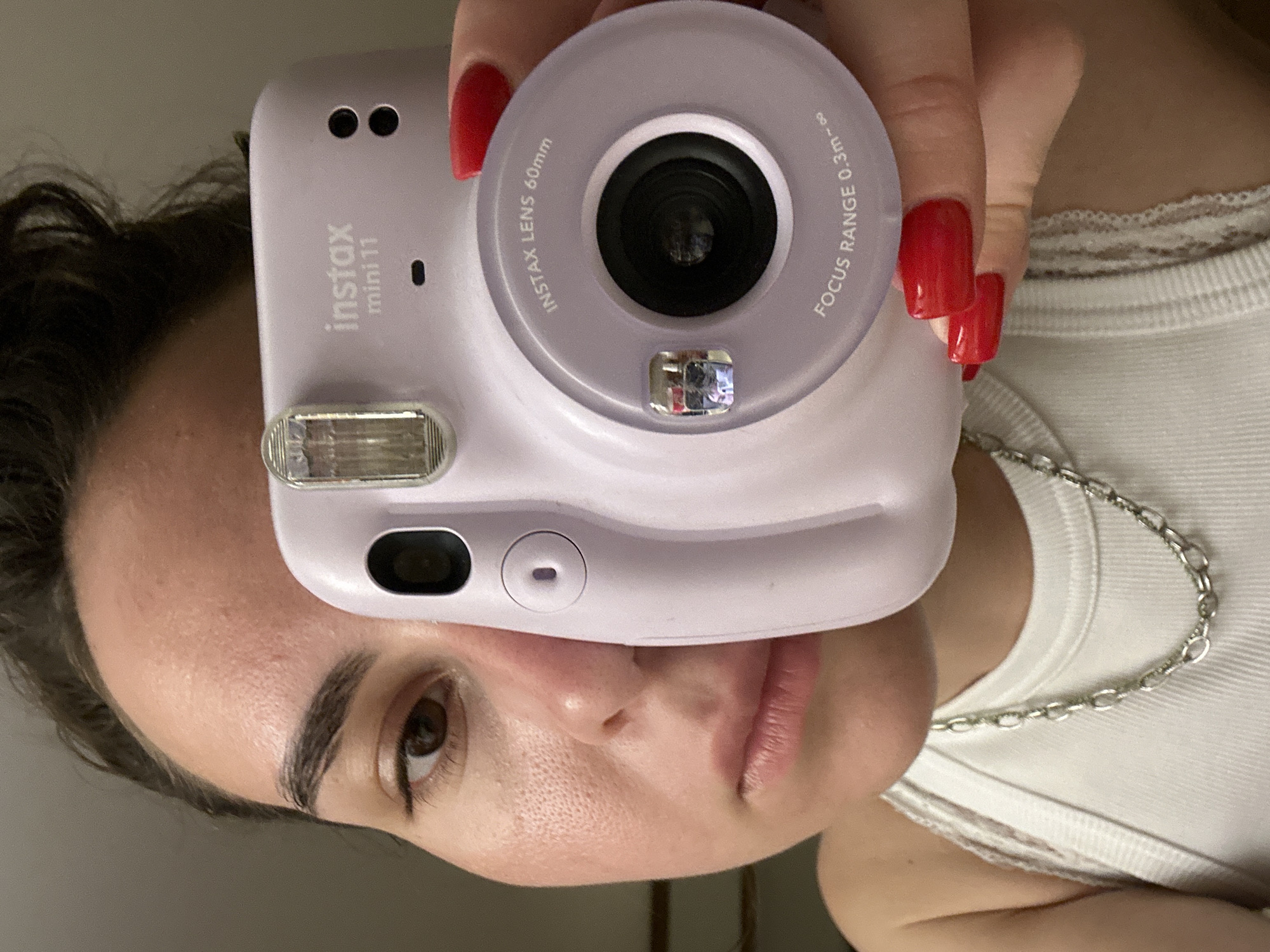 A person holds a purple Instax Mini 11 camera up to their face in an indoor setting. The camera is in the foreground, obscuring part of the face. Red fingernails are visible on the hand holding the camera. The person wears a white tank top and a silver chain necklace. The setting is not identifiable as Pattaya City, Thailand, or any specific location.