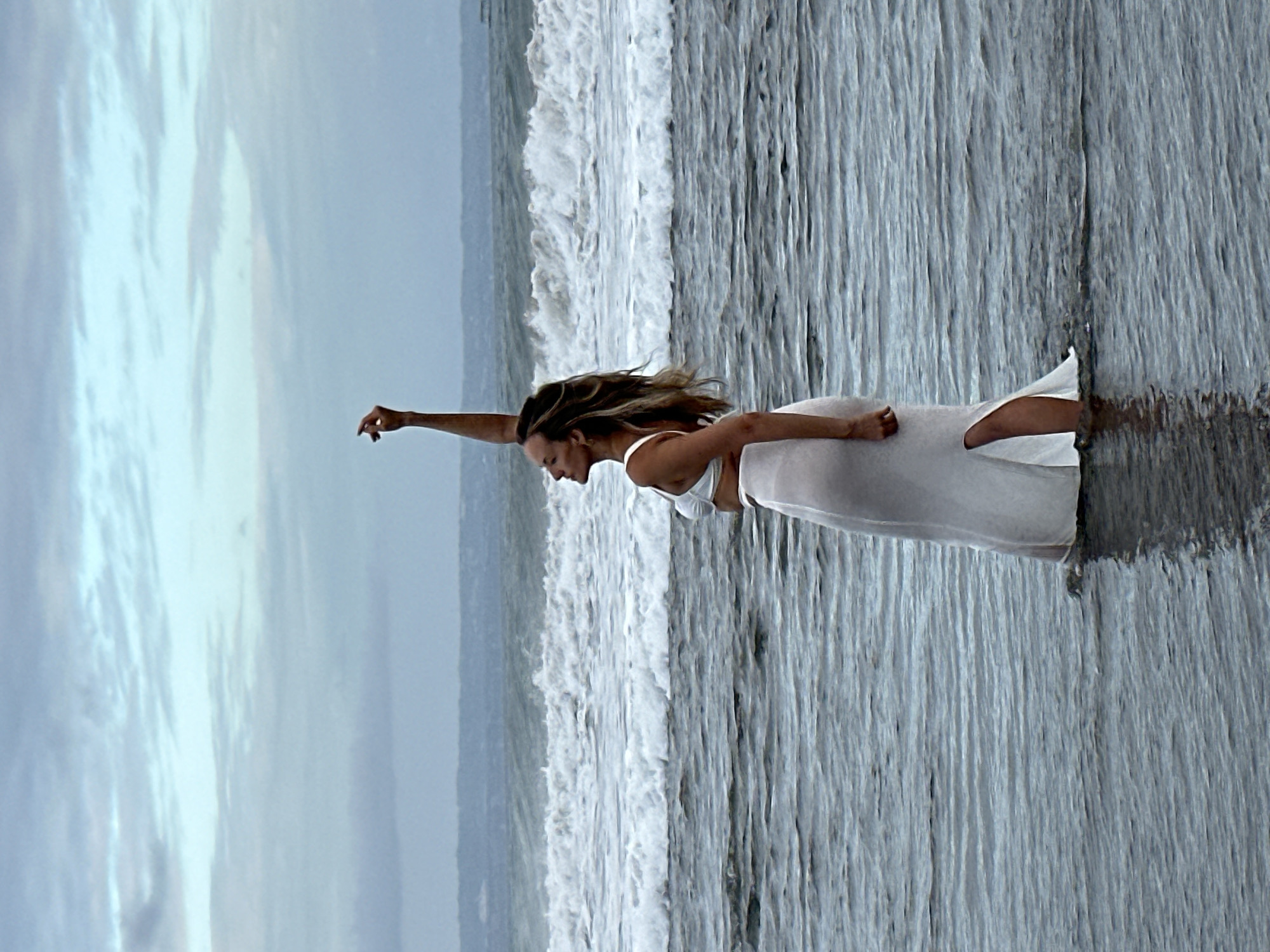 A full shot shows a woman standing in the ocean near North Kuta, Indonesia, with her right arm raised. The woman is wearing a white bikini top and a sheer white skirt with a slit up the side. Her hair is long and wavy, falling around her shoulders. The ocean waves are breaking behind her, and the sky is overcast. The water is a dark gray, and the sand appears to be dark. The image has a somewhat muted color palette, contributing to a serene and slightly moody atmosphere.