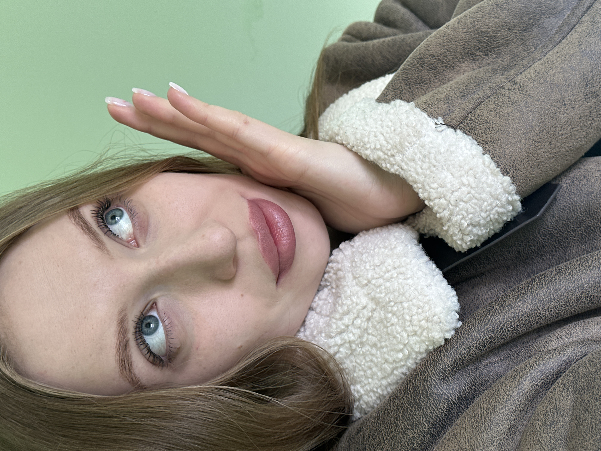 A young woman with light skin, blue eyes, and light brown hair is seen in an image taken indoors. She is wearing a brown jacket with white fleece trim. The location is the city of Chaikovsky, Russia.