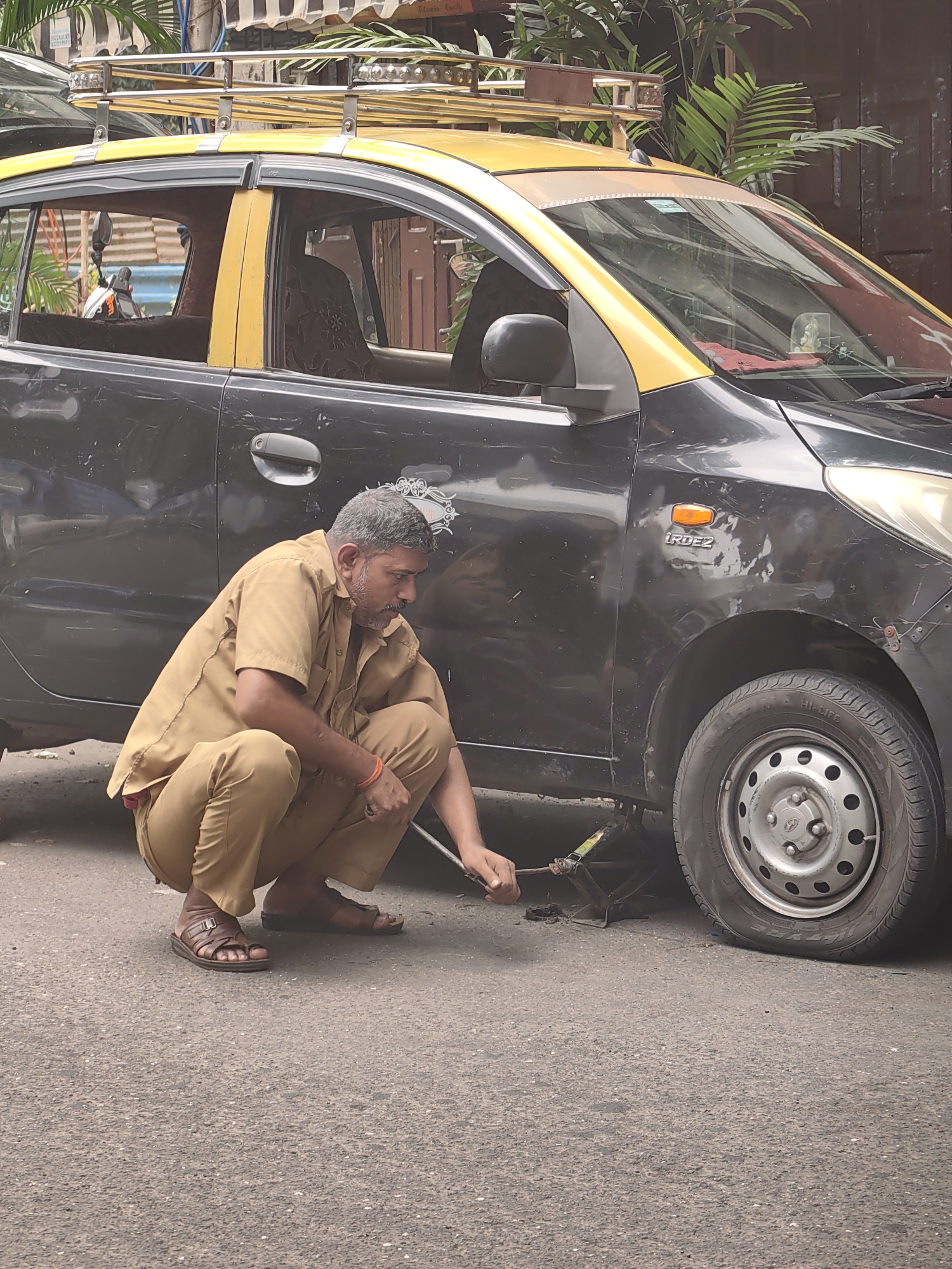 A man, dressed in a beige uniform, is crouching on the asphalt next to a black and yellow taxi. He is operating a car jack, which is positioned under the front passenger side of the vehicle, near the wheel. The jack appears to be lifting the car to change a tire. The taxi has a roof rack with multiple bars. In the background, there are plants and buildings, suggesting an urban environment. The man has a mustache and is wearing sandals. The scene is likely in Mumbai, India, as indicated by the taxi's appearance and the overall streetscape.
