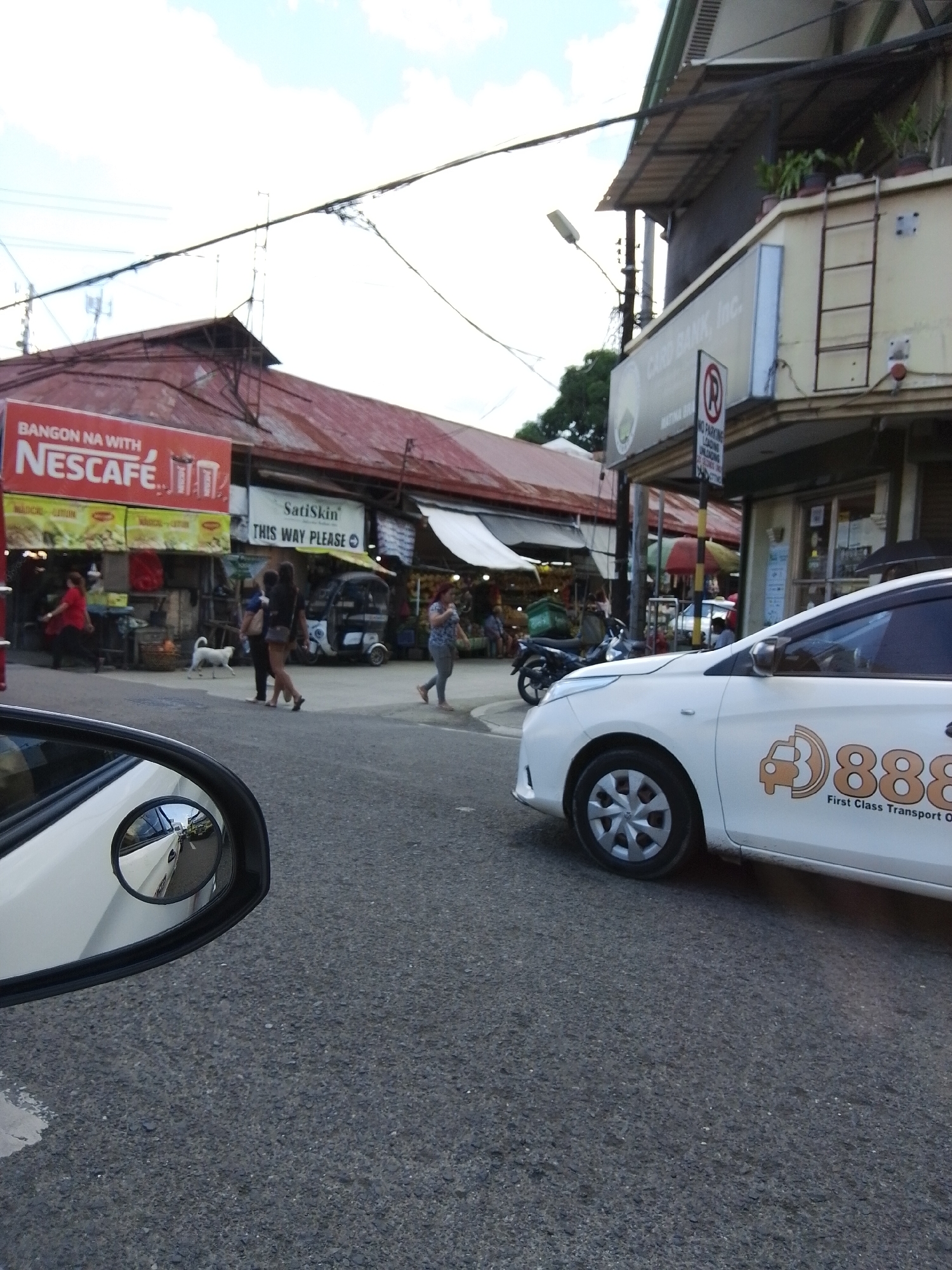 The image captures a street scene in Davao City, Philippines, likely near a commercial area. In the foreground, the side mirror of a white car is visible, reflecting a street with other vehicles. A white taxi with the number "888" and "First Class Transport" written on its side is parked on the right side of the road. Behind the taxi, a building with a sign that reads "CARD BANK, Inc." is visible. Further in the background, there are market stalls with red and white awnings. A sign for "NESCAFÉ" is prominently displayed above one of the stalls. Several people are seen walking in the market area, and a white dog is also present. The sky is partly cloudy. The overall impression is that of a busy urban environment with elements of commerce and everyday life.