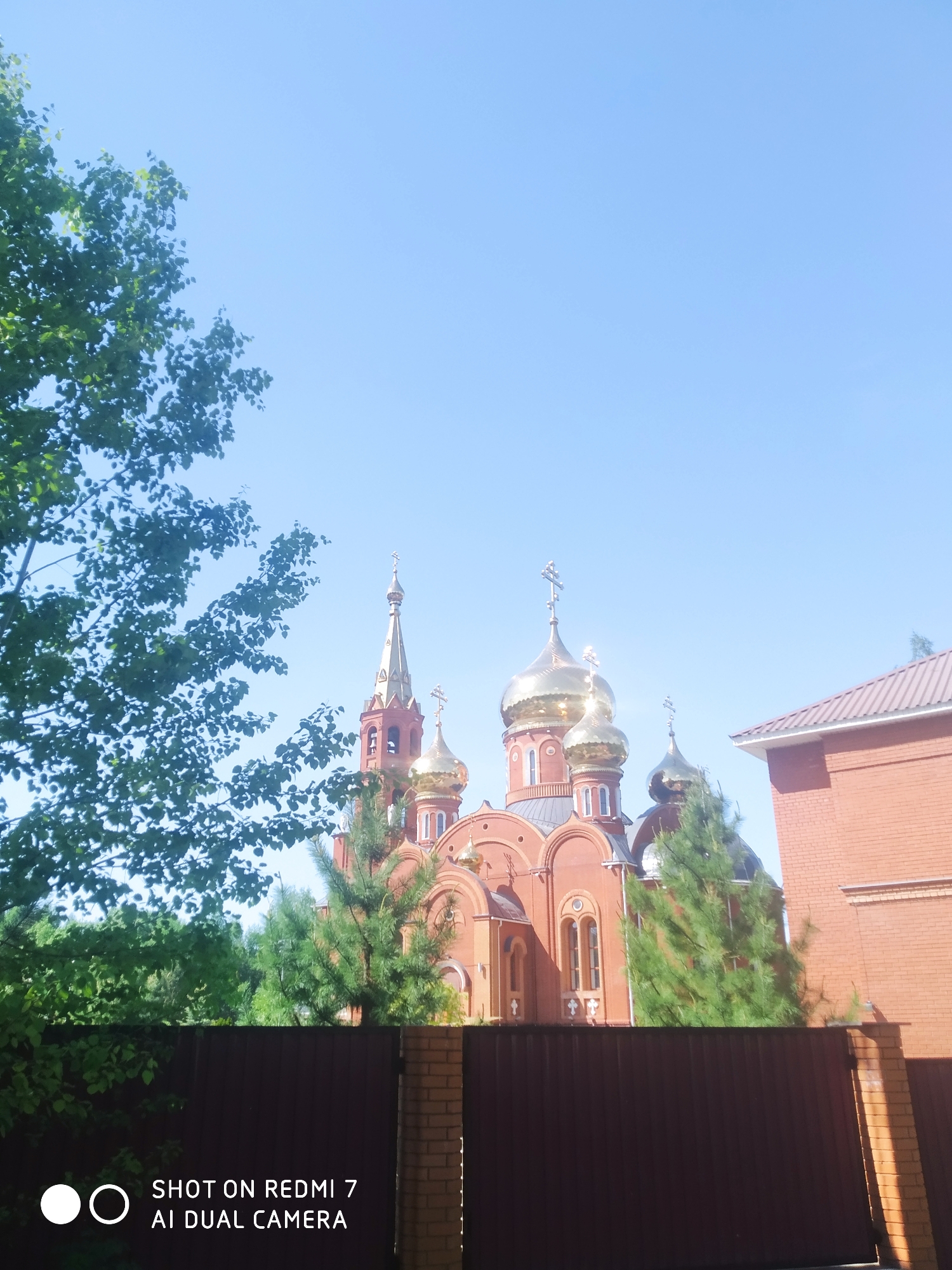 The image shows a red brick Russian Orthodox church with golden domes, located in the city of Chaikovsky, Russia. The church is partially obscured by trees and a dark brown fence. A section of a red brick building is visible to the right of the church. The sky is blue.