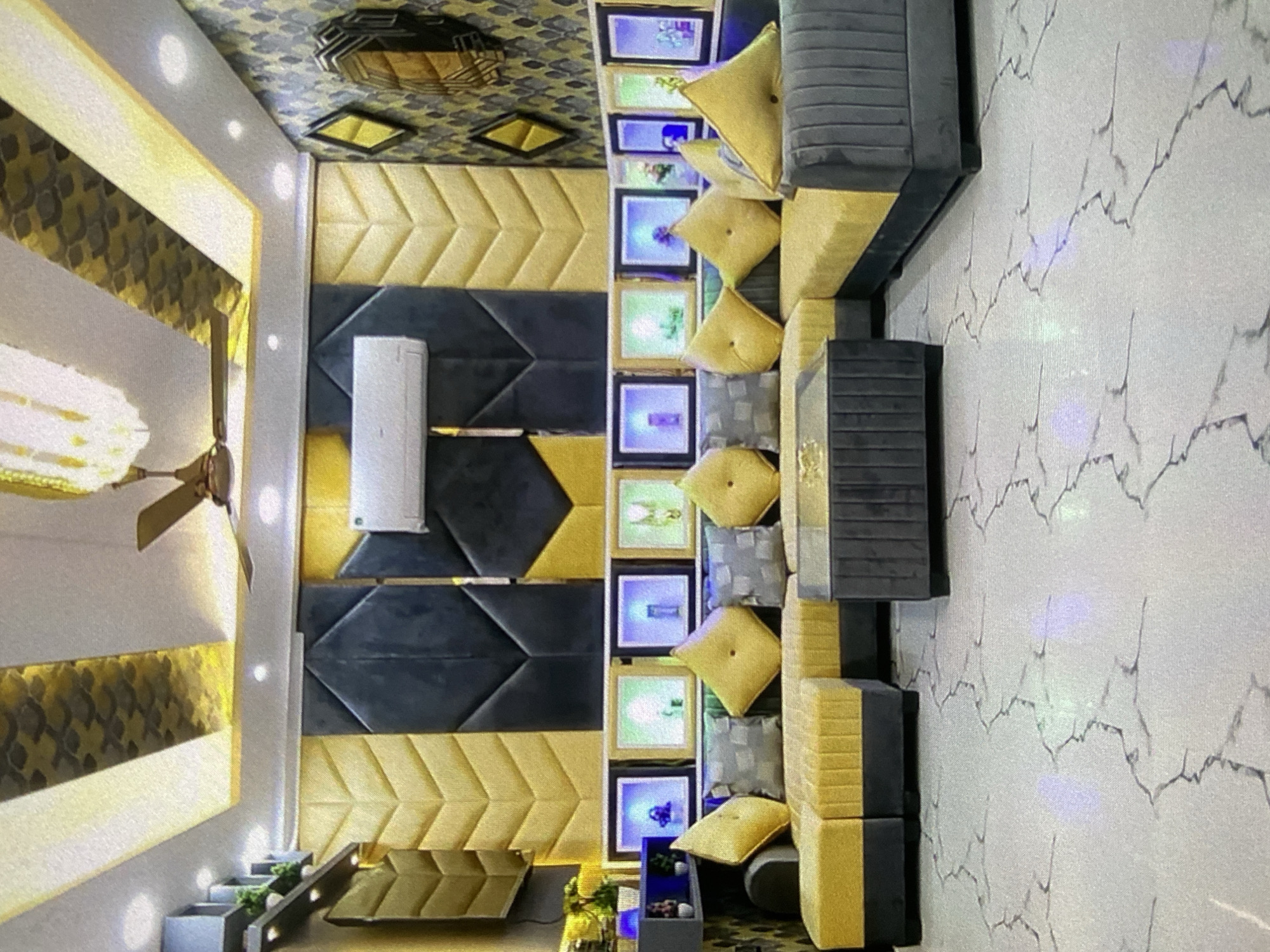 The image shows a room interior with a modern design. The wall is paneled with alternating rows of gold and dark gray chevron patterns. An air conditioning unit is mounted on the wall. There's a display of stacked picture frames between the wall panels and a gray and gold sofa. The sofa has matching yellow pillows. The floor has a white marble pattern. The ceiling has an elaborate gold and black pattern and a ceiling fan. Small potted plants are visible on a ledge near the floor. The location is Lhokseumawe, Indonesia.