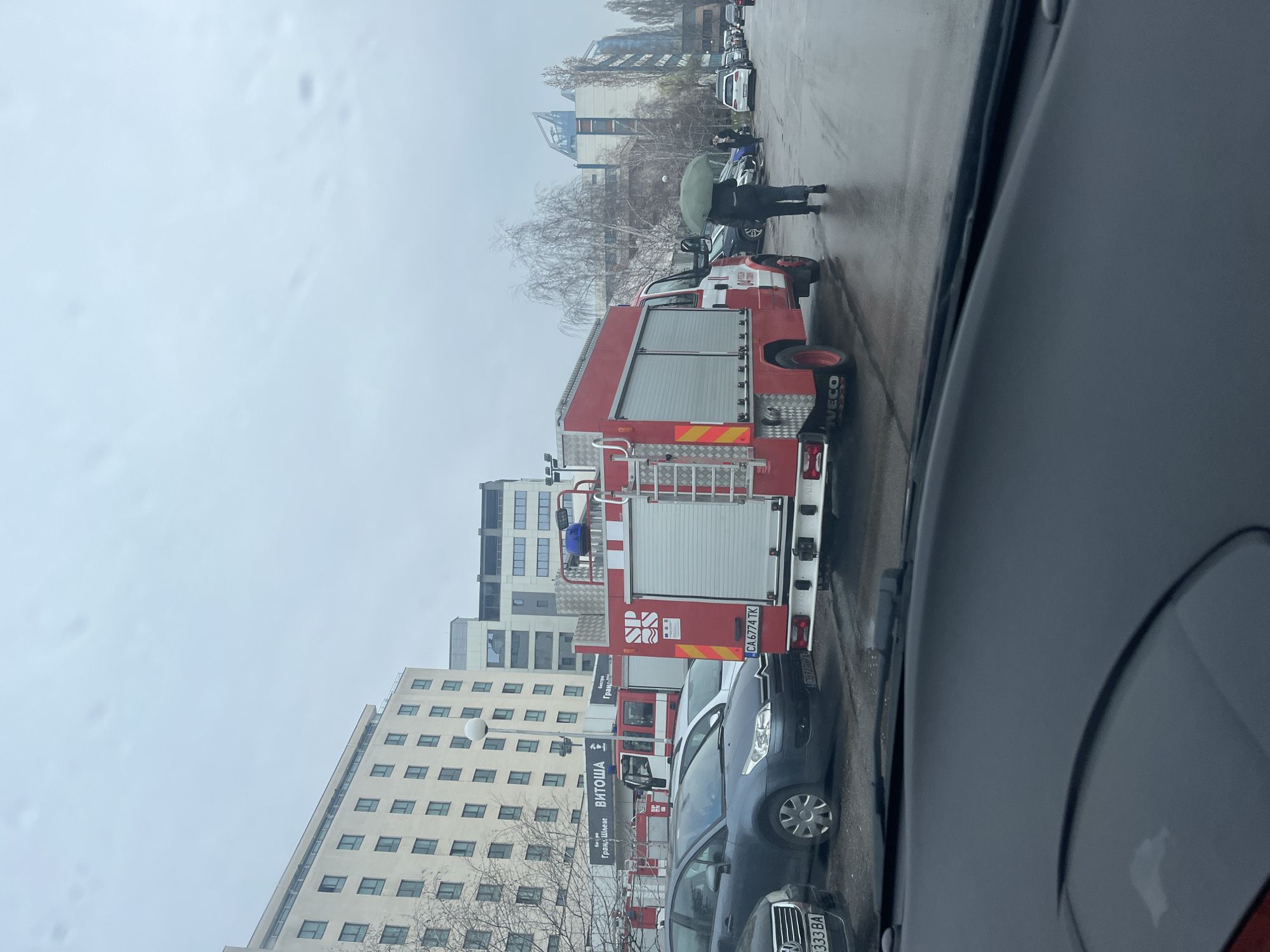 The image depicts a street scene in Sofia, Bulgaria, on a cloudy day. A red fire engine is parked on the side of the road, partially obstructing the view. Other parked cars are visible in the foreground and background. A person with an umbrella is walking on the sidewalk near the fire engine. Modern buildings are visible across the street. "Vitosha" is visible on one of the buildings. The photo was taken from inside a car, as part of the car's interior is visible in the foreground. The license plate of the fire truck is CA 6774 TK.