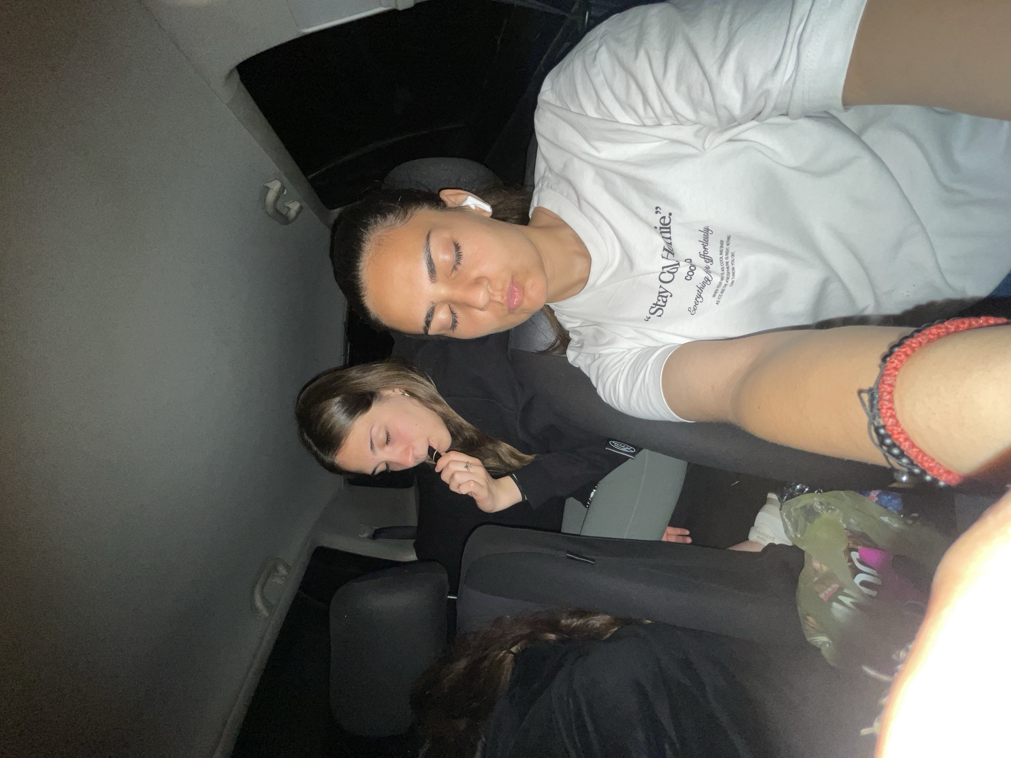 The image shows three young women in a car. The car's interior is dark, suggesting it is night or a poorly lit area. The woman closest to the camera is wearing a white t-shirt with the words "Stay Co Homie" printed on it. She appears to be sleeping or resting her eyes. An AirPod is visible in her ear. Another woman is sitting behind her, wearing a black top and is awake, perhaps holding something near her mouth. Part of a third woman is visible at the bottom of the frame, wearing a black jacket. A plastic bag containing items like a white New Balance shoe is visible near the bottom of the frame. There's a red and black beaded bracelet on the wrist of the person taking the picture. The interior of the car is grey.
Based on the prompt information, it is not possible to confirm any location.