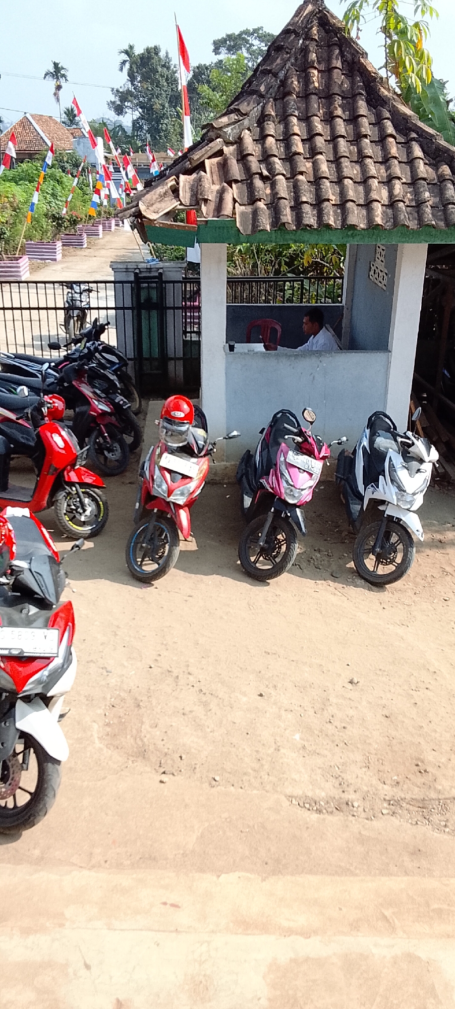 In the city of Tanjung Setia, Indonesia, a row of parked scooters is visible, with some adorned with red and white flags, suggesting a national celebration. The scooters are of various colors, including red, pink, and white, and some have helmets on them. Behind the scooters, a small building with a tiled roof stands, and a man in a white shirt is seated inside. The ground is covered in dirt and gravel. The background shows trees, houses, and more flags. The image captures a street scene, likely in a residential or public area, during a time of festivity.
