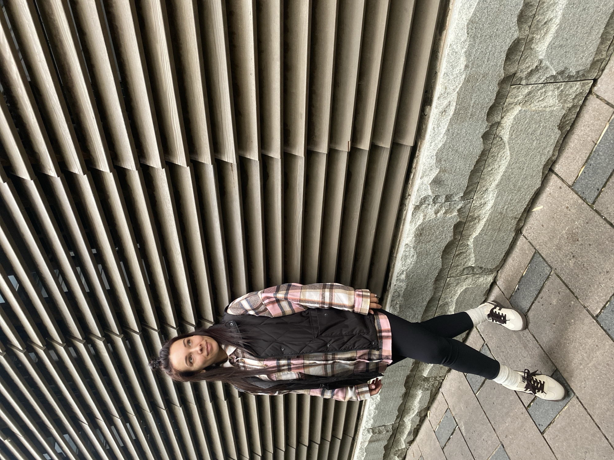 A woman stands outdoors against a building with a slatted facade and a stone foundation. She has long, dark hair and wears a black vest over a pink and white plaid shirt, black leggings, and white sneakers with black laces. She faces the camera with a slight smile. The background consists of the textured slats of the building on one side and the stone foundation transitioning into a paved walkway on the other. The location is not identifiable as Krasnodar, Russia from the image alone.