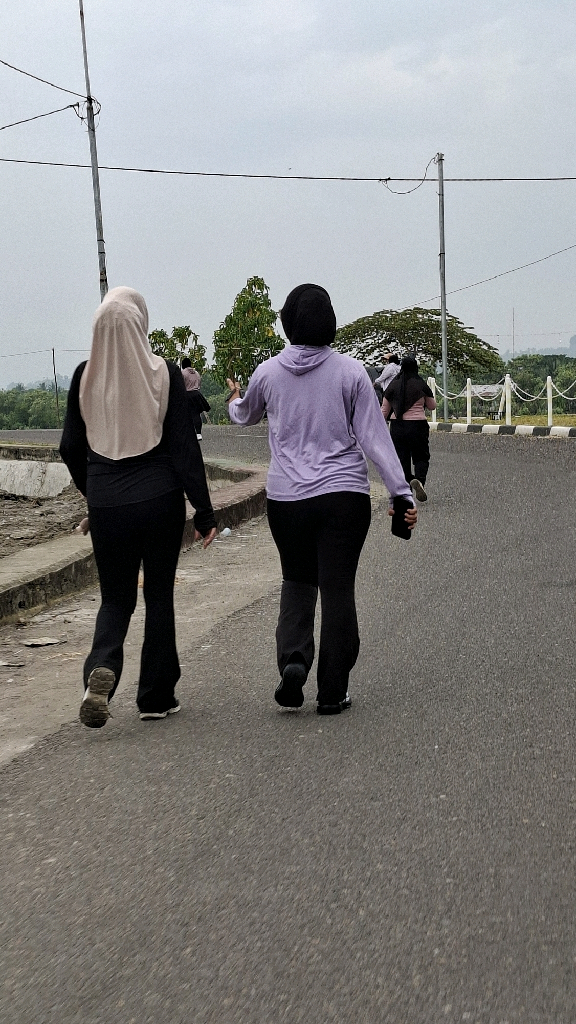 The image depicts an outdoor scene, likely in Lhokseumawe, Indonesia, under an overcast sky. In the foreground, two women are walking away from the camera on an asphalt road. The woman on the left wears a light-colored hijab and black athletic wear, while the woman on the right wears a dark hijab and a lavender hooded sweatshirt with black pants. The woman on the right is holding a dark object, possibly a phone, in her right hand. Further ahead, two more individuals, dressed in dark athletic wear, are also walking on the road. To the left of the road, there is a concrete barrier with some debris. In the background, trees and a white fence with black accents are visible, suggesting a park or recreational area. Utility poles and overhead wires are present in the upper portion of the image.