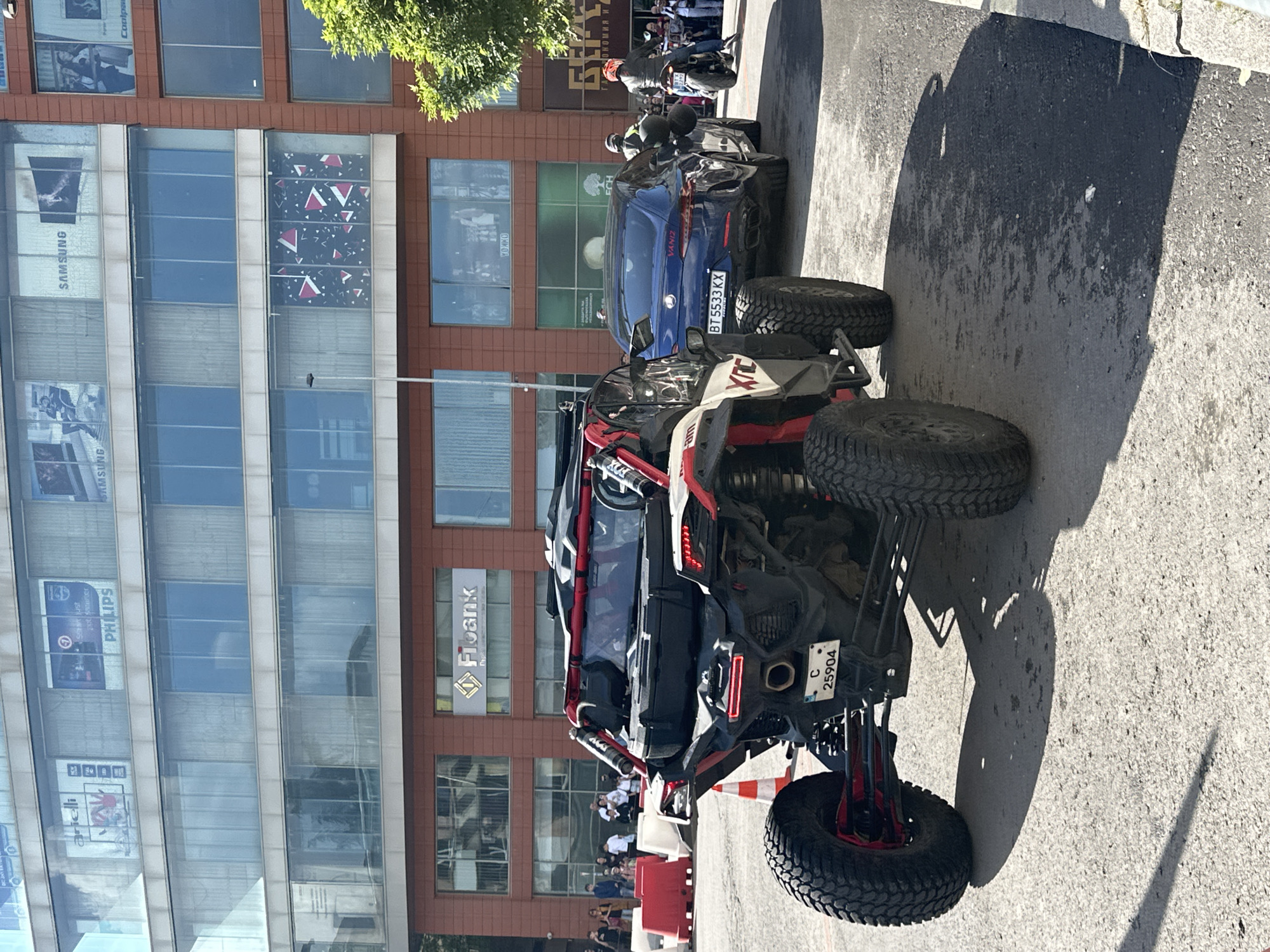 The image shows an off-road vehicle parked on a street with a modern building in the background. The vehicle is black and red, with large tires. A blue car is parked behind it, and a motorcycle is visible further back. People are standing near the sidewalk, observing the scene. Advertisements for Samsung and Philips are visible on the building. A sign for "Fibank" is also present. The license plate of the blue car reads "BT5533 KX," and the off-road vehicle has a license plate "C 25904." The location appears to be in Veliko Tarnovo, Bulgaria.