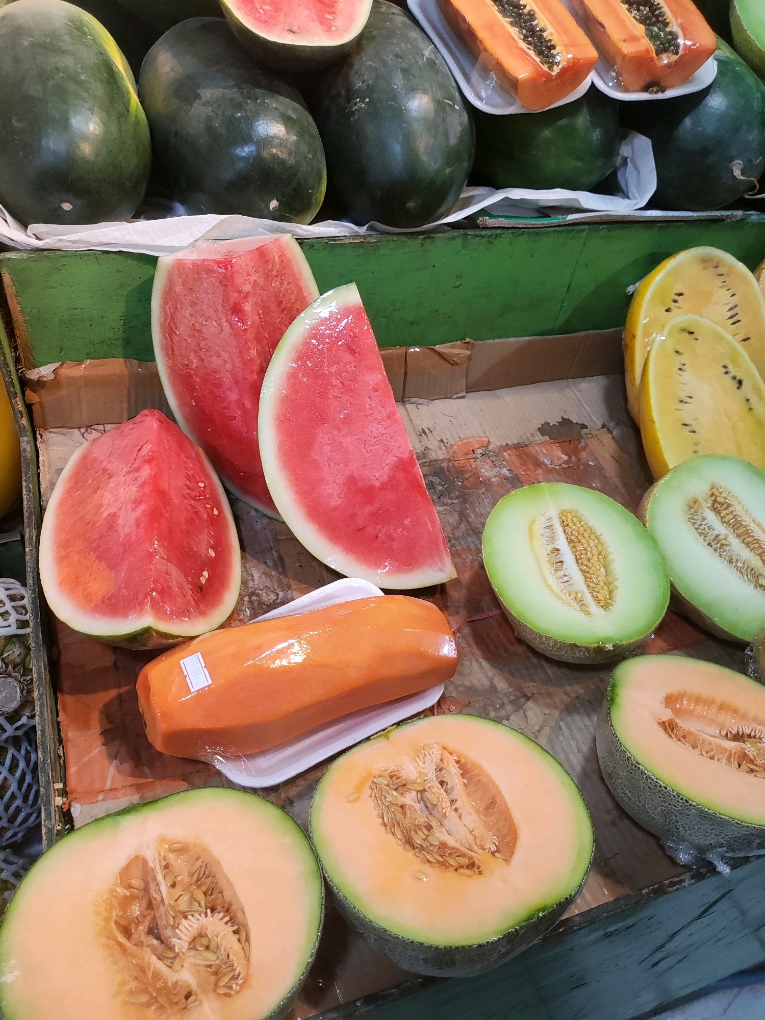 A variety of fresh fruits are displayed in a market stall. In the foreground, two halved cantaloupe melons with bright orange flesh and visible seeds are laid out. To the left of these, a large, whole watermelon is cut into a wedge, revealing its red, juicy interior. Next to this, a single, whole papaya, wrapped in plastic, sits on a white tray. Behind these, more watermelon wedges are visible, along with whole watermelons in the background. The stall also features yellow melons and other fruits that are partially obscured. The scene is illuminated by overhead lighting, creating natural shadows and highlights on the fruits. The market stall appears to be located in Telagaasih, Indonesia.