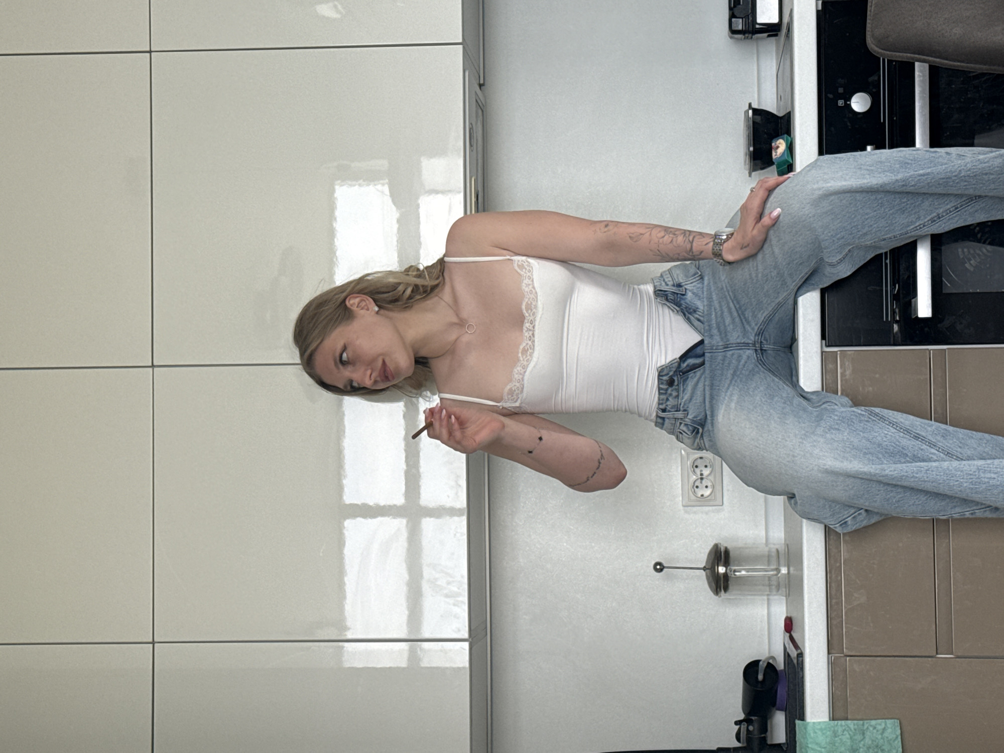 A woman stands in a kitchen with white cabinets in Rostov-on-Don, Russia. She wears a white tank top, blue jeans, and holds a rolled cigarette. A white outlet is seen on the kitchen wall.