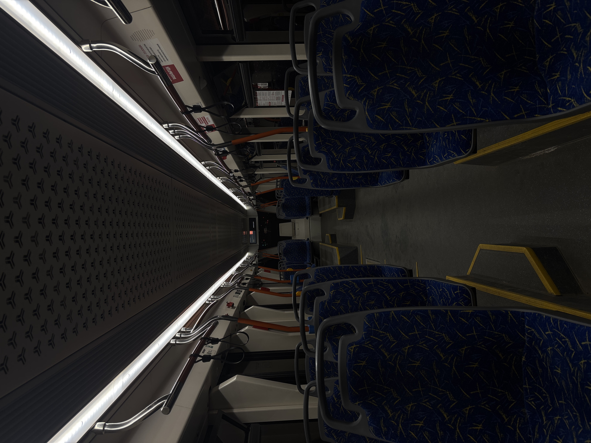 Here's a detailed description of the image:
**Scene:** The interior of a public transport vehicle, likely a bus or tram, in Perm, Russia.
**Environment:** The image presents a long, narrow aisle flanked by rows of seats. The lighting is predominantly artificial, with a bright, linear light fixture running along the ceiling on the left side. The floor appears dark.
**Objects:**
* Rows of seats with blue and yellow patterned upholstery.
* Handrails and grab bars extending from the ceiling or side panels, presumably for standing passengers.
* Wall panels with a repeating geometric pattern.
* Signage, possibly containing safety information or advertisements (some in Russian).
**Additional Detail:** The seats appear unoccupied.
