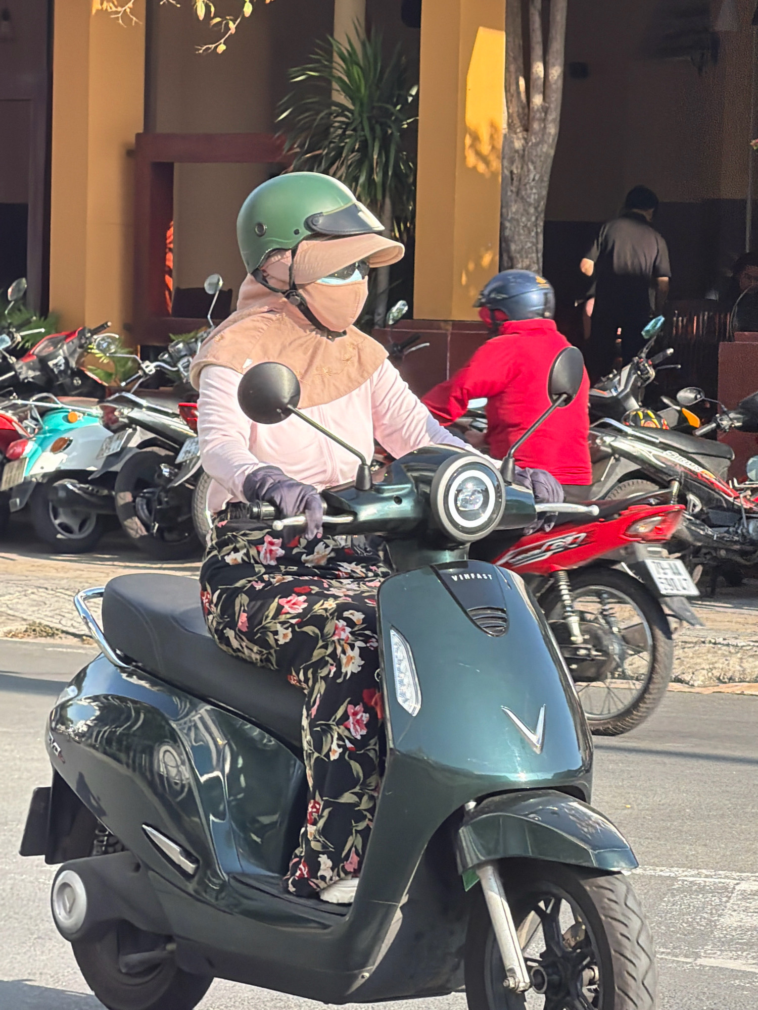 A person wearing a green helmet, light pink shirt, dark patterned pants, and a face mask is riding a dark green scooter. The scooter has a "VINFAST" logo on its front. The person is also wearing dark gloves and a beige head covering. In the background, other motorbikes are parked, and a person in a red jacket is visible on another scooter. The scene appears to be on a street in Nha Trang, Vietnam, indicated by the style of the scooters and the surrounding buildings. The lighting suggests it is daytime, with sunlight casting shadows.