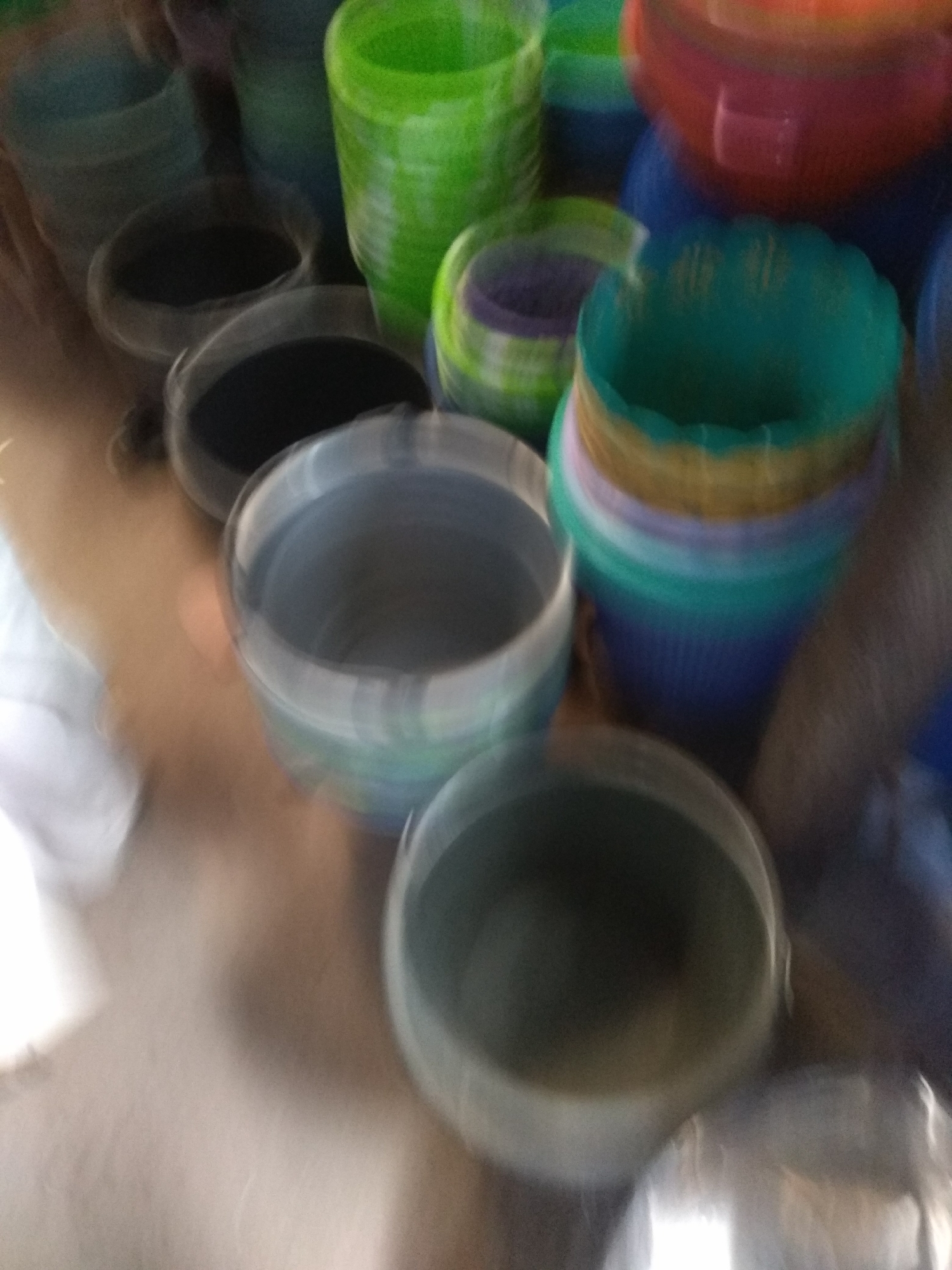 The image depicts a collection of various colored plastic containers, possibly buckets or pots, arranged in a somewhat haphazard manner. The containers are in different sizes and colors, including green, blue, red, and grey. Some are stacked, while others are placed next to each other. The scene appears to be an indoor setting, possibly a shop or storage area. The lighting is diffused, and the image is slightly blurred, suggesting movement or low light conditions. No people are clearly visible in the image. The arrangement of the containers does not provide specific clues about their intended use beyond general storage or display. The background is indistinct and appears to be a plain wall or surface.