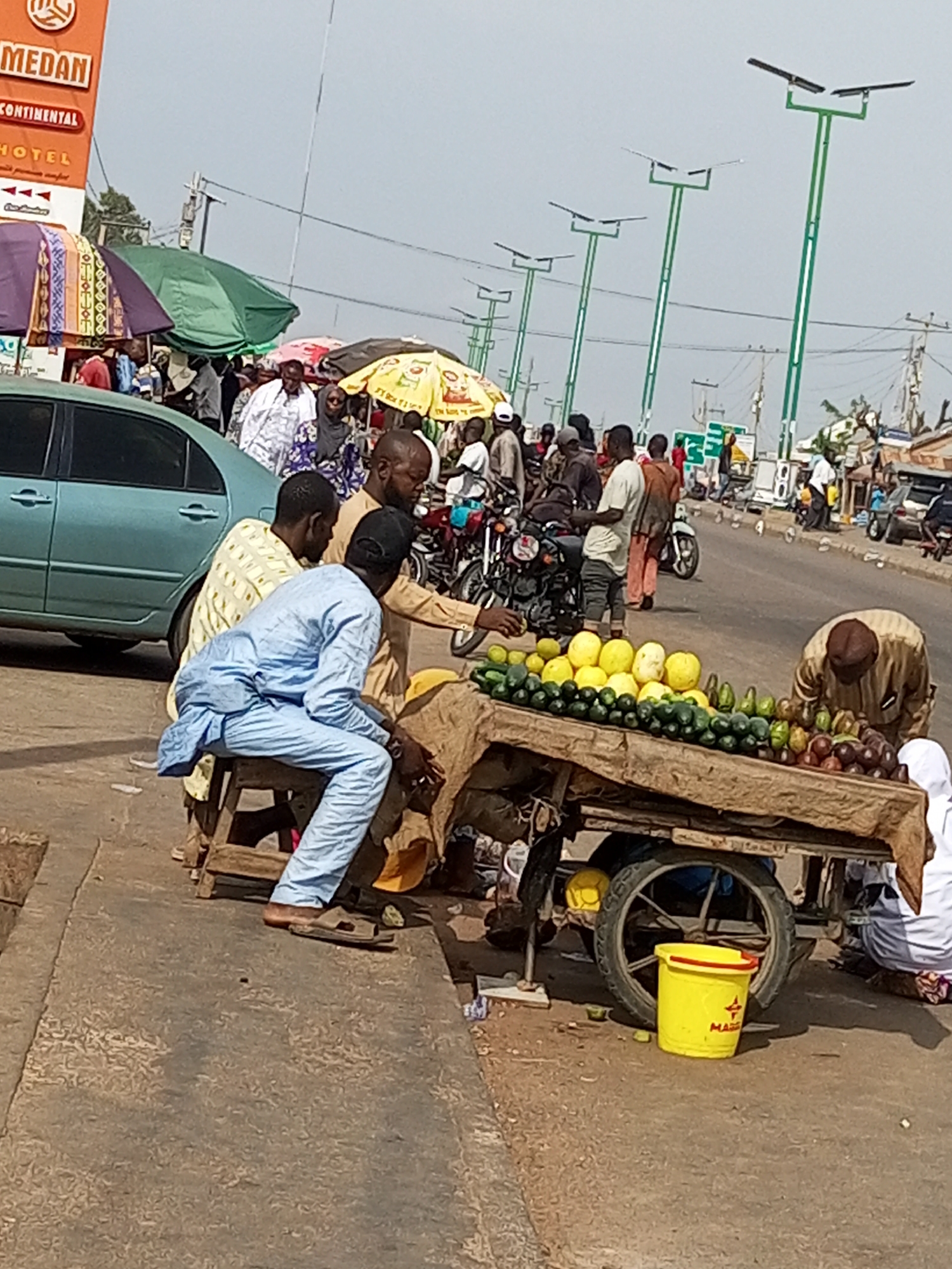 An outdoor street scene shows a fruit and vegetable cart being tended to by a vendor. The cart is laden with produce including cucumbers, avocados, and what appear to be yellow melons or citrus fruits. A yellow bucket sits near the cart. Several people are gathered around the cart, some appearing to be customers and others the vendor and his associates. In the background, there are parked vehicles, motorcycles, and a street lined with vendors under umbrellas and awnings. Several modern streetlights are visible in the distance. A sign for "MEDAN CONTINENTAL HOTEL" is also visible.