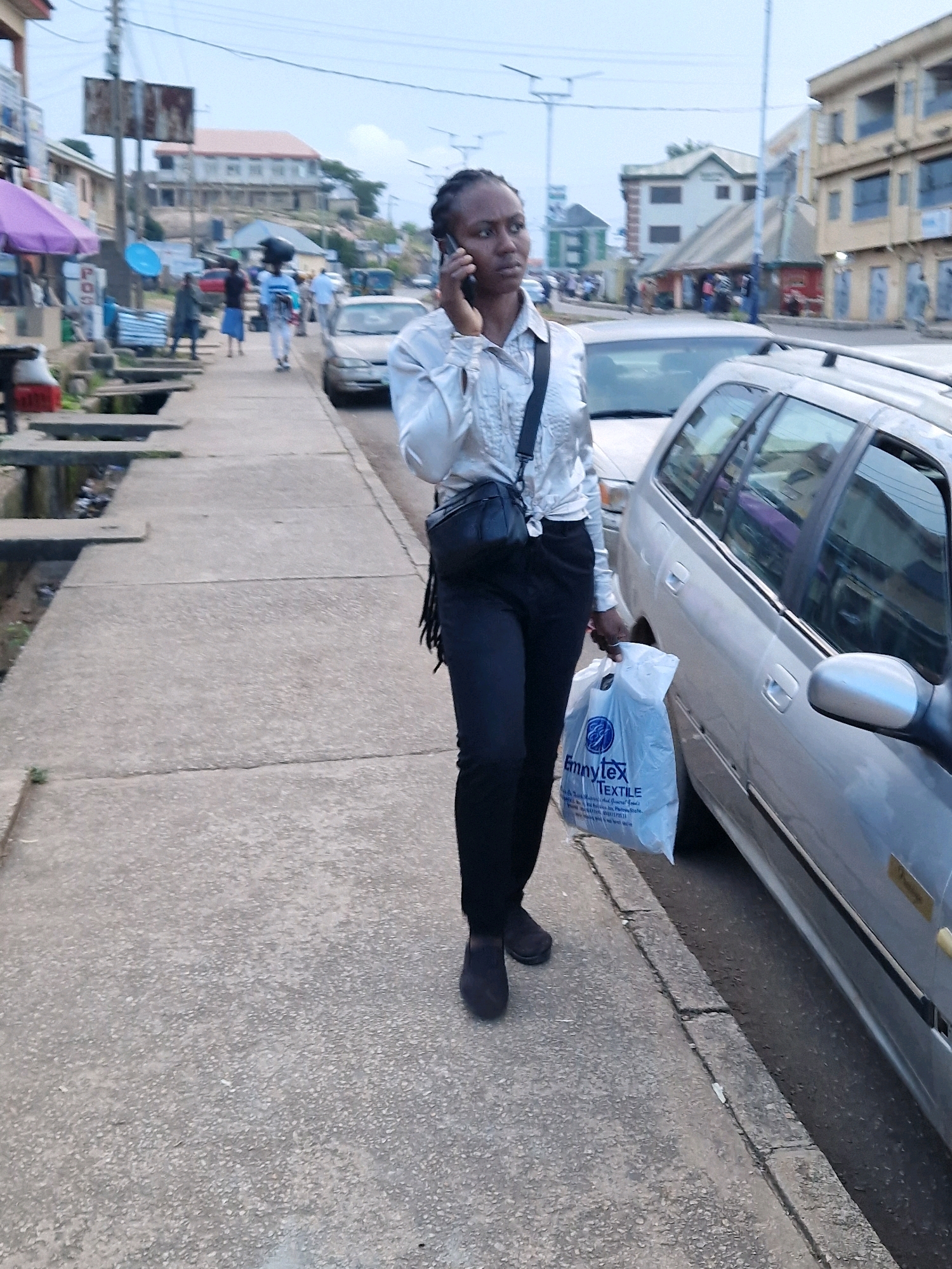 In Kabong, Nigeria, a woman walks along a concrete sidewalk next to a parked silver station wagon, talking on her cell phone and carrying a white plastic shopping bag. She is dressed in a long-sleeved, light-colored, shimmering blouse and black pants, with a black crossbody bag over her shoulder. The shopping bag she carries has a logo that reads "Emmytex TEXTILE" and some text below it in smaller font. The environment suggests a street with buildings lining one side, including what appears to be shops or residences with awnings and balconies. The sky is overcast, indicating a possibly humid or rainy day. The street itself is paved, and the sidewalk is segmented by expansion joints. Other individuals and vehicles are visible in the background, suggesting this is a moderately busy urban area.