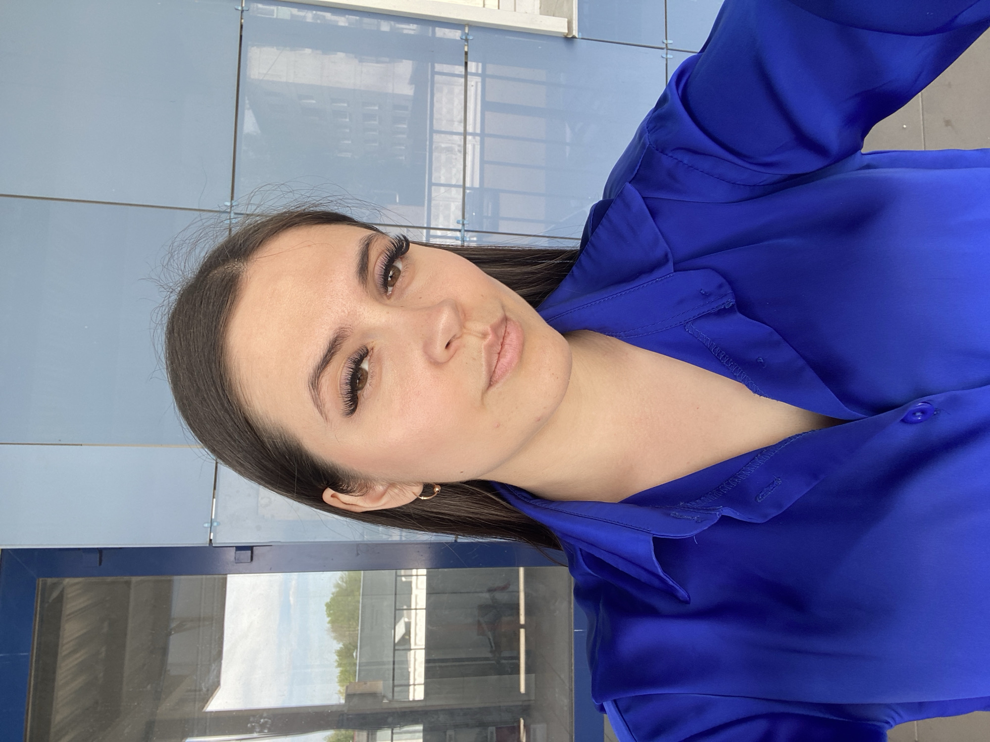 A woman with dark hair and fair skin is facing the camera, wearing a bright blue blouse. She appears to be standing near a glass facade that reflects a cityscape. The location is indicated as the city of Chaikovsky, Russia, though specific features of the location are not discernible in the image.