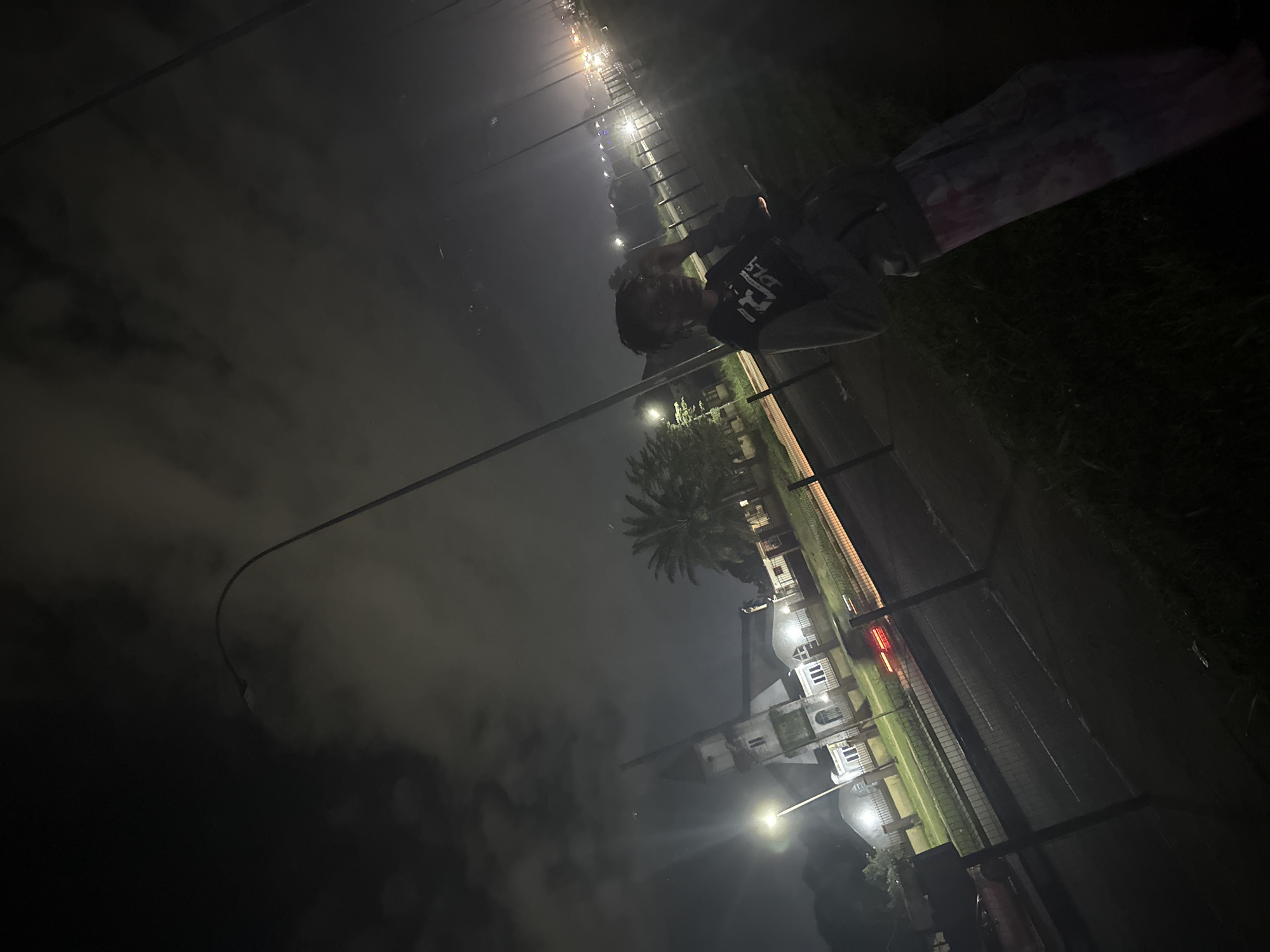 A person stands in the foreground to the right of the image, facing slightly away from the camera. They are wearing a gray hoodie over a black t-shirt and tie-dye pants. Their hair is dark and styled in braids. In the background, a large white building with a steeple, possibly a church, is illuminated by streetlights. The building is set back from a road with a fence in front of it. Palm trees are visible behind the building. The scene is at night, with streetlights casting a glow and clouds obscuring the sky. The location is the city of Uyo, Nigeria.