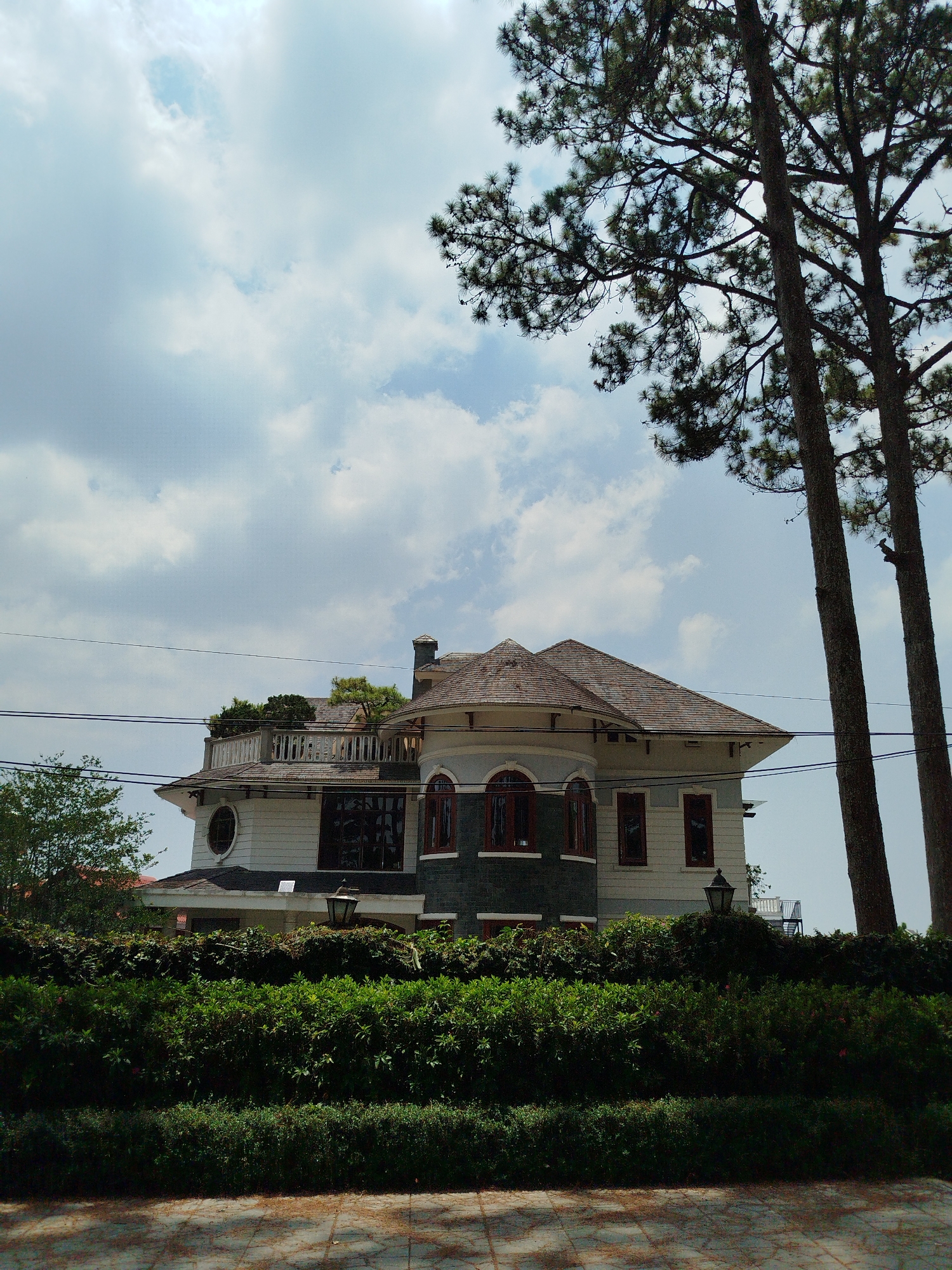 The image shows a two-story building in what appears to be Da Lat, Vietnam, based on architectural style and vegetation. The building has a brown tiled roof, light-colored walls, and a partially visible balcony on the second floor. Some windows are rounded and trimmed in red. A chimney is visible. There are green plants on top of the balcony, and a dark gray brick section at the base of one of the building's sections. Two tall, slender trees stand to the right of the building. A dense green hedge or shrubbery partially obscures the lower section of the building. A weathered stone or paved surface is in the foreground. Power lines run across the sky above the building. The sky is light blue with white clouds. Two black lampposts are visible.