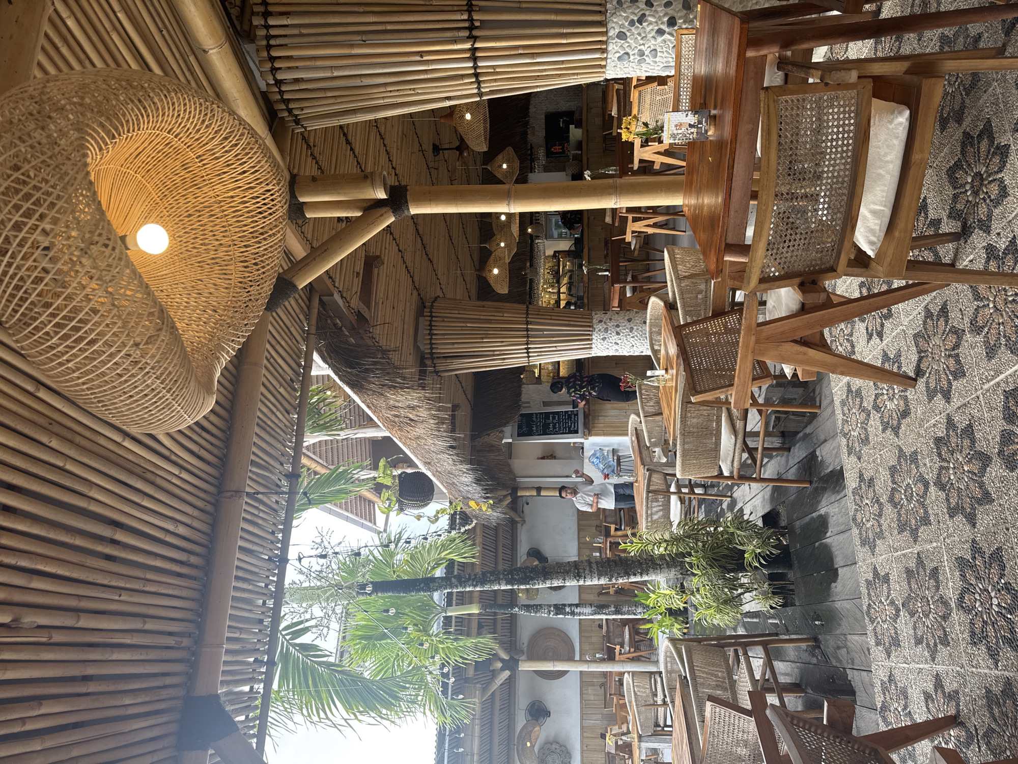 This image captures the interior of a restaurant in Nusa Penida, Indonesia. The structure features a prominent use of bamboo in its construction. The roofing and walls are made primarily of bamboo, with woven elements. A large, decorative woven lamp hangs from the ceiling. The seating area comprises wooden tables and chairs with woven backs. The flooring is patterned with a floral design created from small stones. People can be seen inside, including staff and a customer taking a photo. Outside, a palm tree is visible, contributing to the tropical ambiance.