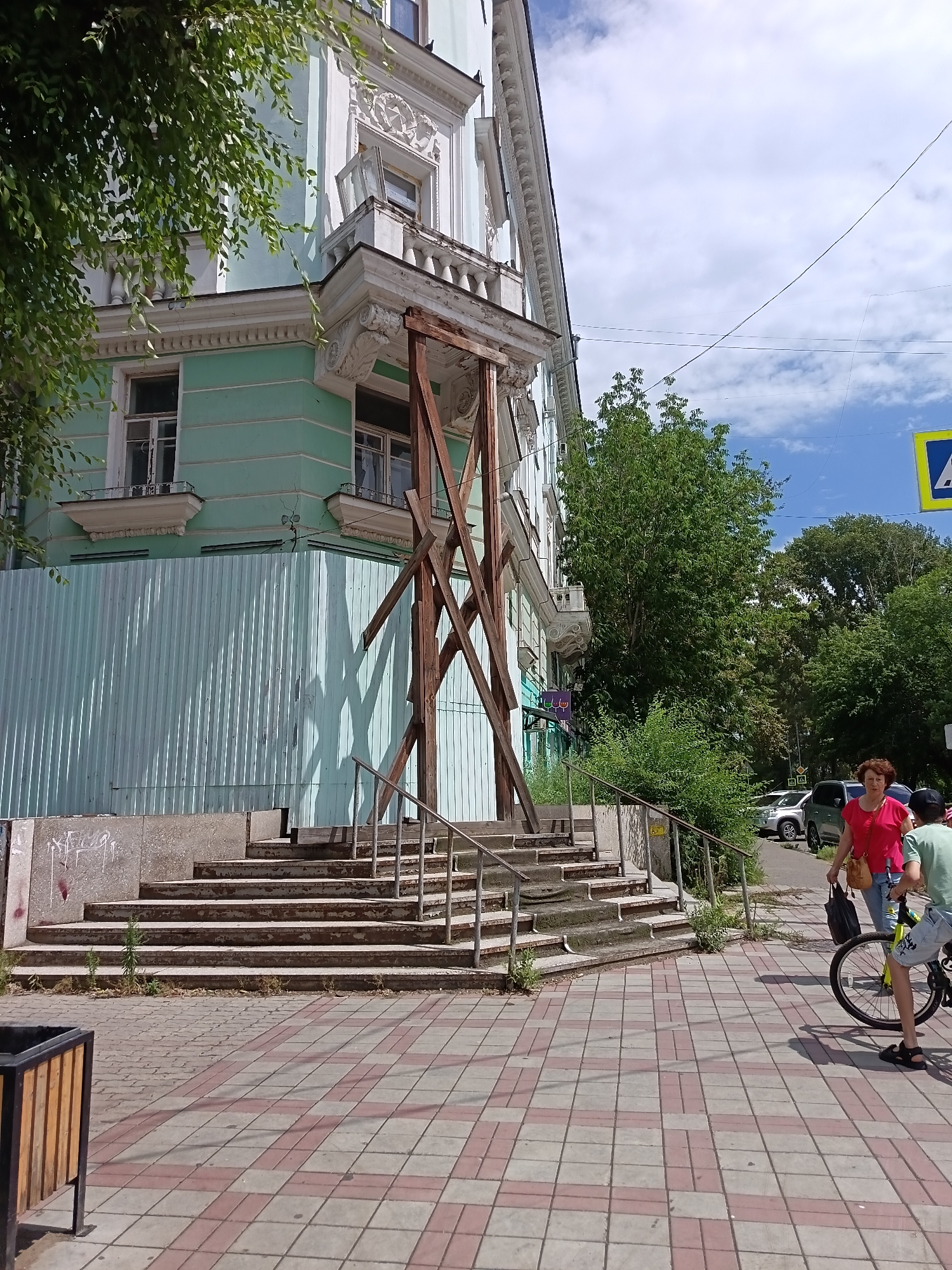 A medium shot shows the exterior of a light blue and white ornate building with a wooden scaffolding structure in front of its entrance steps. The building has multiple windows, balconies with decorative railings, and cornices. A tall fence made of corrugated metal panels is erected in front of the lower part of the building, obscuring a portion of the entrance. The scaffolding, constructed from rough-hewn wooden beams, is positioned on the right side of the steps, leaning against the building and extending upwards. The steps themselves are made of stone and appear weathered, with some vegetation growing around their base. A metal handrail runs along the side of the steps.
In the foreground, a paved walkway with a pattern of light and dark tiles runs horizontally. To the right of the steps, a woman in a bright pink shirt and jeans walks towards the viewer, carrying a dark bag. Next to her, a person on a bicycle, wearing a light green t-shirt and denim shorts, is also facing the viewer. The bicycle is yellow-green. Further in the background, cars are parked along a street lined with trees. The sky is blue with scattered clouds. The scene appears to be in an urban environment. The location is the city of Komsomolsk-on-Amur, Russia.