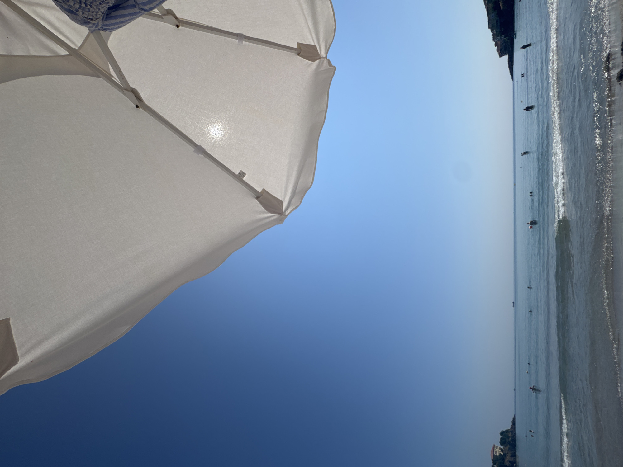 The image depicts a sunny day at a beach in Sozopol, Bulgaria. The foreground is dominated by the underside of a white beach umbrella, casting a shadow and revealing the umbrella's frame and a striped blue and white towel draped over it. The bright blue sky fills the upper portion of the image.
The middle ground features the calm sea with several people swimming and wading. Gentle waves are breaking on the shore. In the distance, a few boats are visible on the horizon.
The background shows the coastline of Sozopol. On the left, a building with a red roof is perched on a rocky outcrop. On the right, a rugged cliff face is visible. The overall scene suggests a relaxed summer day at the seaside.