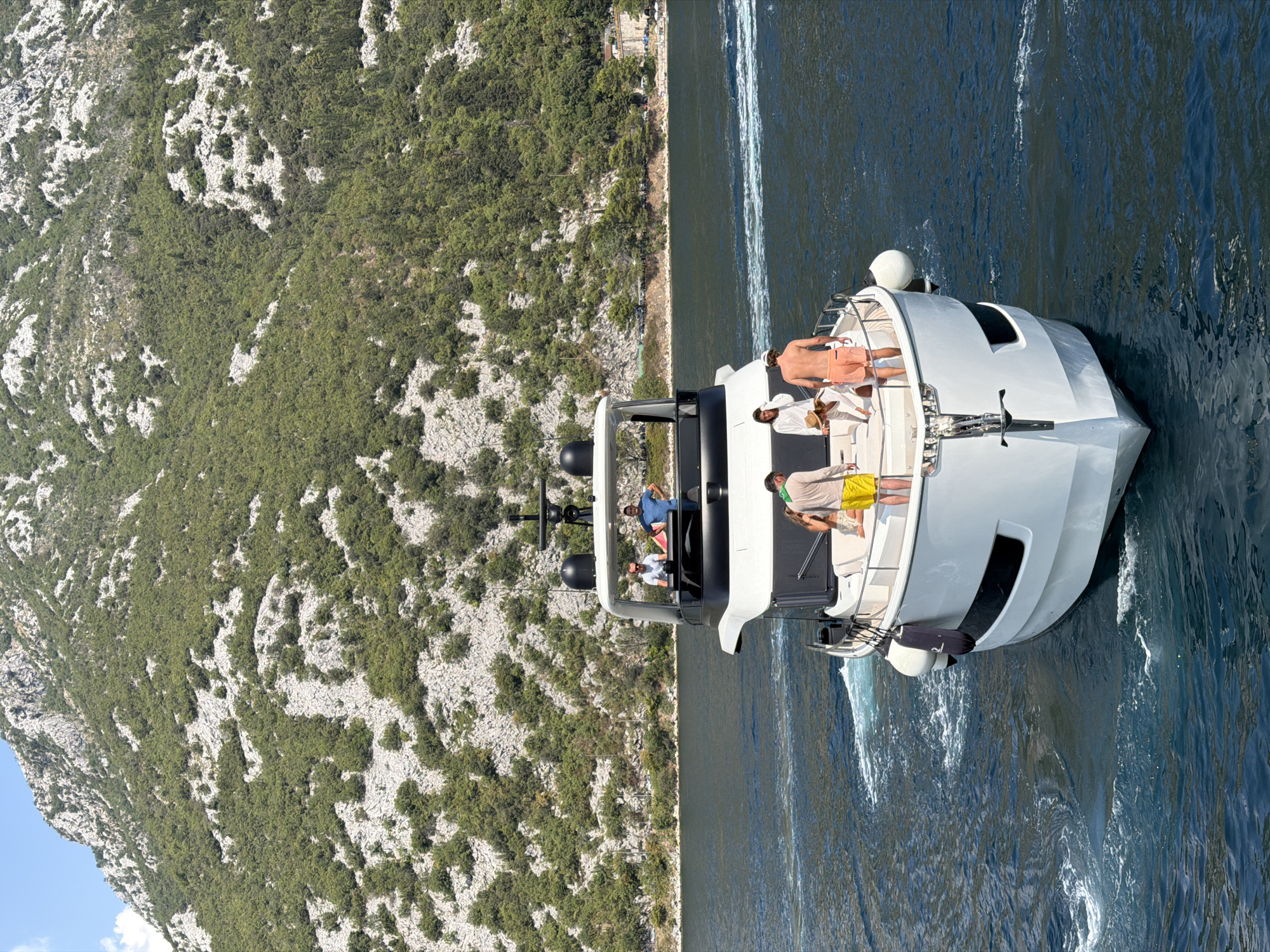 In the Bay of Kotor, near the town of Perast, Montenegro, a white luxury yacht is navigating through choppy, dark blue water, leaving a foamy wake. The yacht has a sleek, modern design with multiple levels. On the upper deck, two men in casual wear, one in a blue t-shirt and the other in a white polo shirt, stand near the helm. On the lower deck, at the stern, four individuals are visible. A man in yellow shorts and a light-colored shirt stands with his back to the camera, while a person in a white dress and straw hat is leaning down. Another man in orange shorts is also present. Behind them, a woman with long dark hair, wearing a light-colored top and patterned shorts, is seated. The yacht is framed against a backdrop of a steep, rocky mountainside covered in green vegetation. The sky is clear and blue, with a few wispy white clouds.