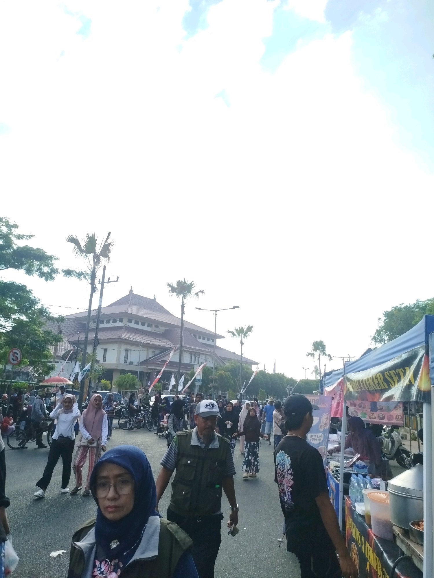 The image depicts a street scene in Bangkalan, Indonesia, likely a public event or market. In the foreground, a woman wearing a blue hijab and glasses is looking towards the camera. To her right, a man in a green vest and a white cap is walking away. Further back, a crowd of people is milling about, some walking, others standing. There are several stalls set up along the side of the street, with awnings and goods displayed. One stall appears to be selling food, with bottles of water and containers of ingredients visible. Behind the stalls, palm trees and a building with a distinctive roof are visible. The sky is bright and cloudy, suggesting it is daytime. The overall atmosphere appears lively and bustling.